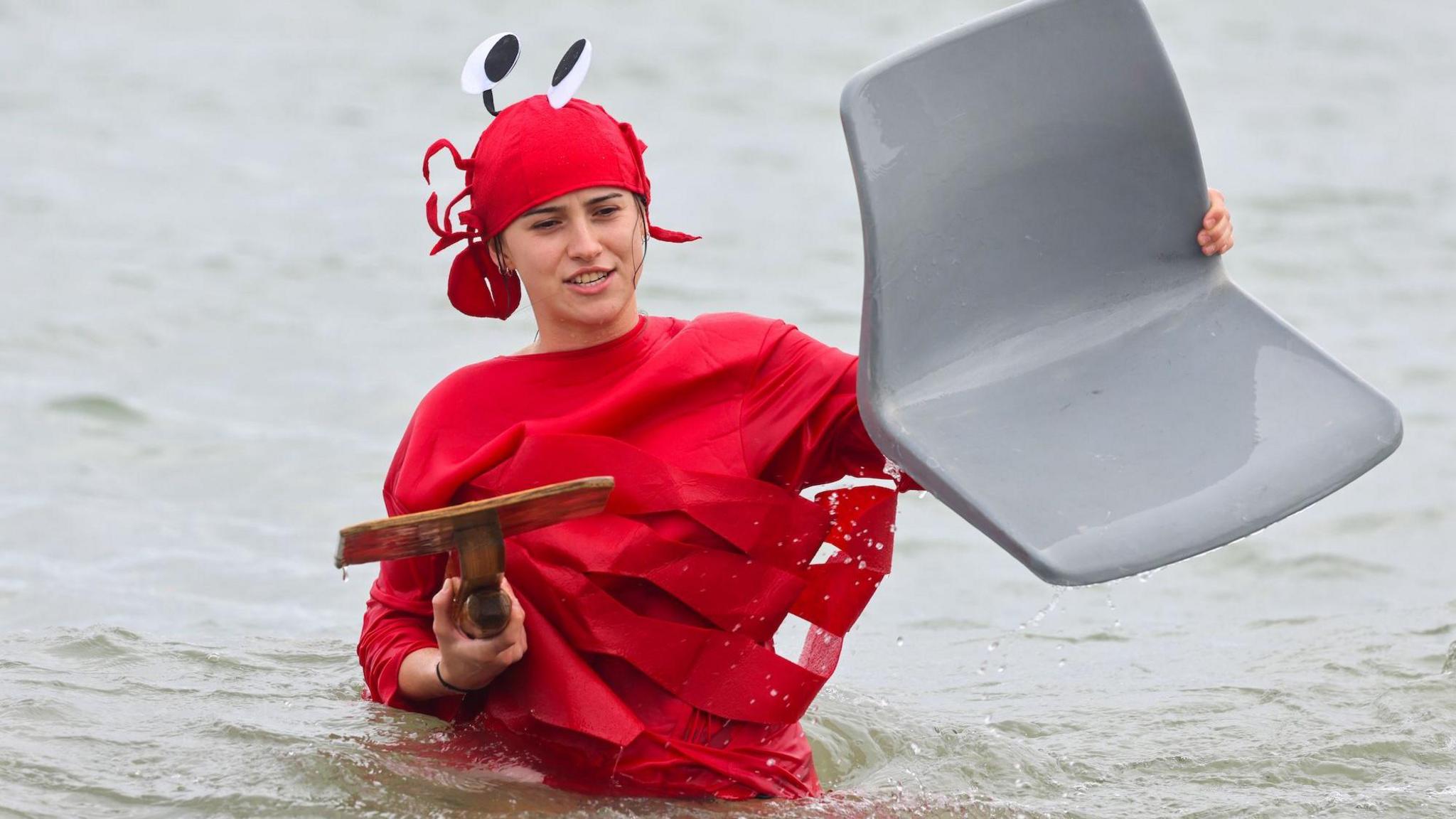 A woman in a lobster costume stands in the seat, holding a bit of wood and plastic seat.