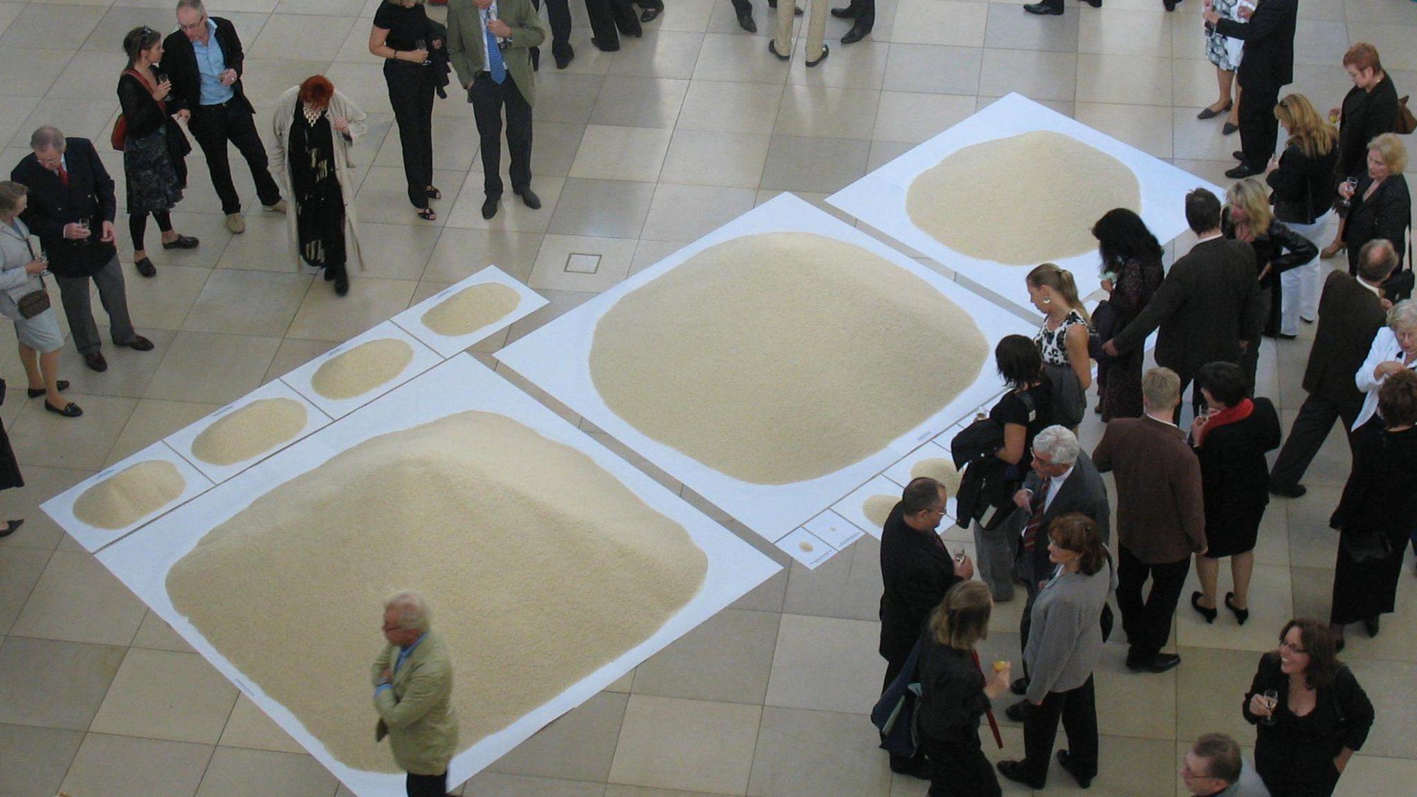 There are three large white squares placed diagonally across the floor, each with different amounts of rice piled on top of them. The first on the left has the most rice piled up, and it goes down in descending order. Above the first square are four rectangles in a line next to each other with similar amounts of rice on them. There are people gathered around either side of the displays, some talking to one another and others looking on at the piles