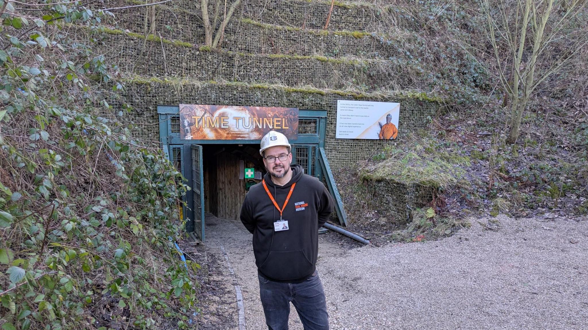 A man wearing a hard hat and black tracksuit top stands in front of a sign reading 'Time tunnel'