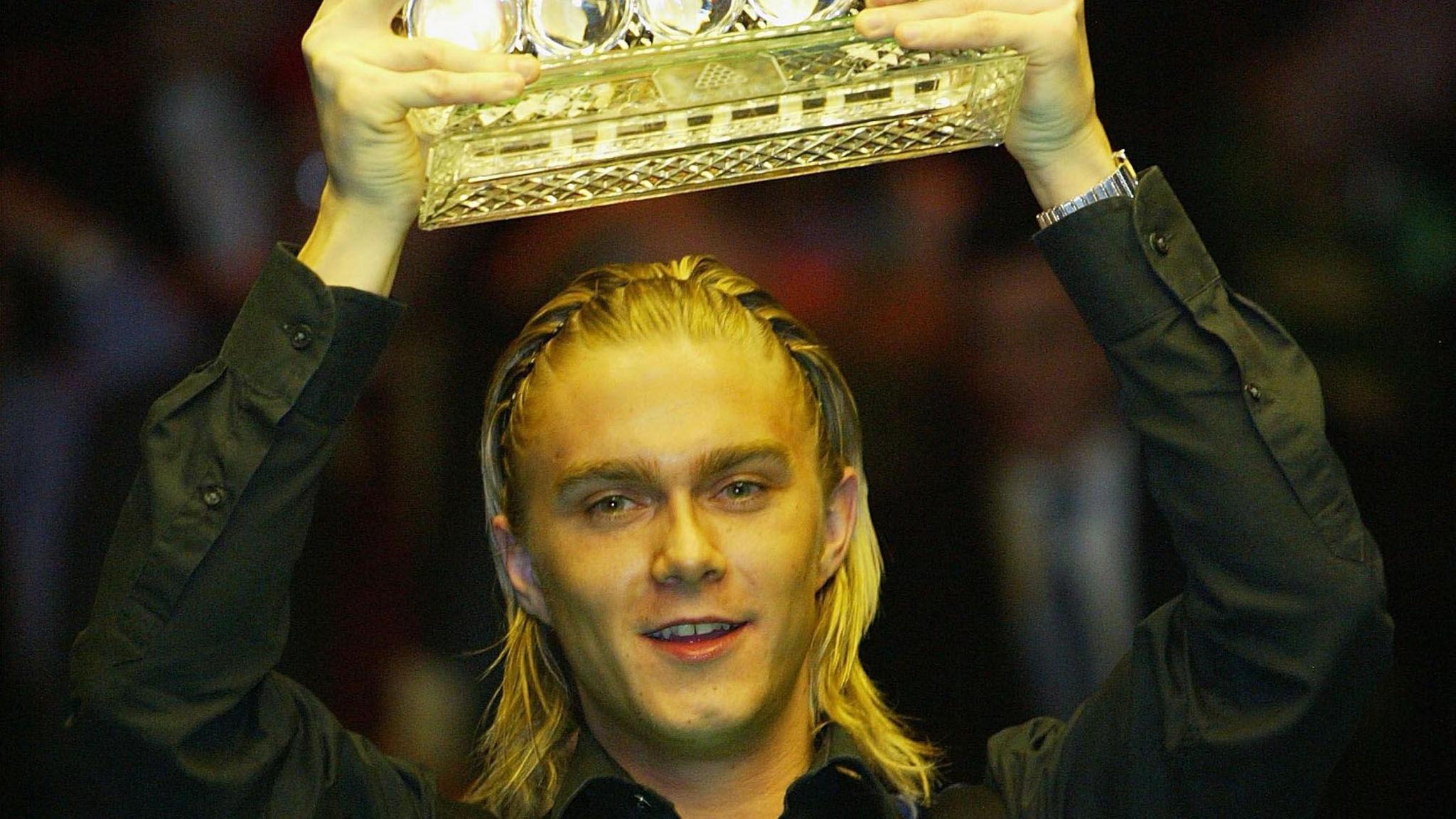 The snooker player Paul Hunter, lifting a trophy aloft out of shot. His blond hair is braided and he is wearing a black shirt.