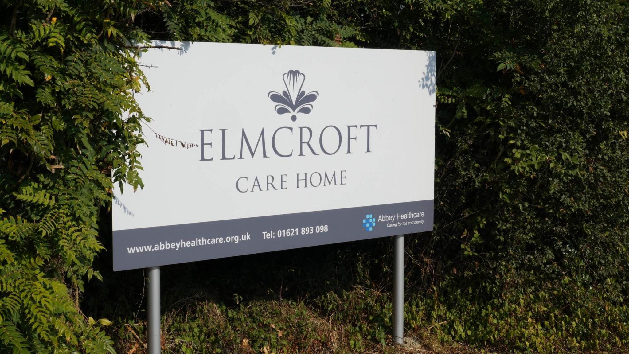 A sign at the entrance of Elmcroft Care Home. It says the home's name and provides a contact number and website. The sign itself is white and has grey lettering.