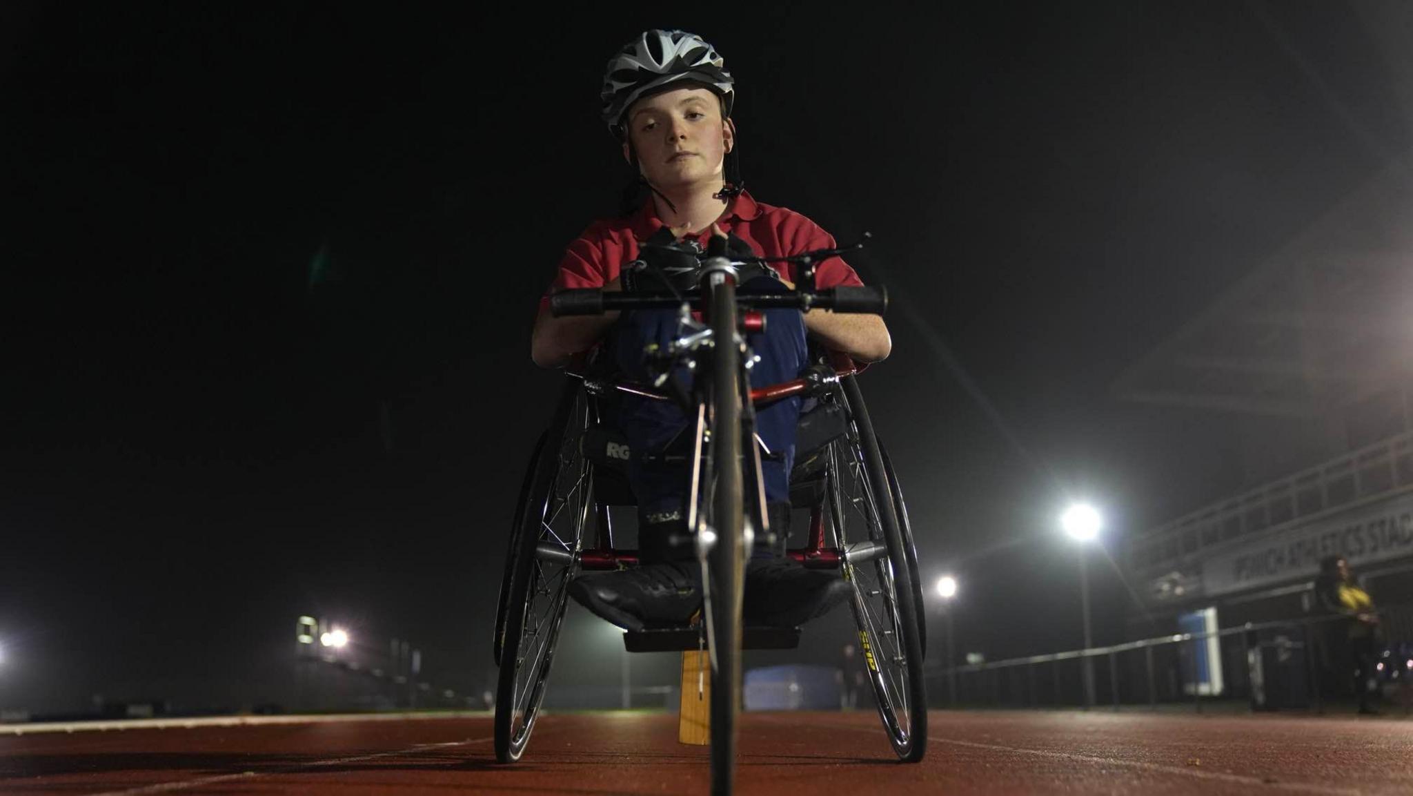 Morgan is pictured in his racing wheelchair on the athletics track. He is looking down at the camera with a determined expression on his face. He is wearing a silver helmet and a red t-shirt with racing gloves on his hand.