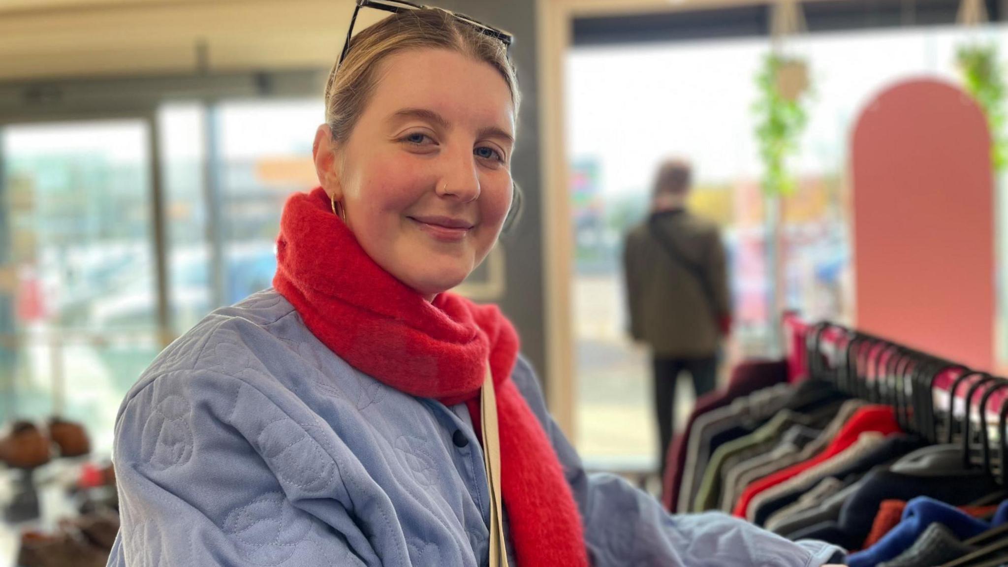 A young woman with a blue coat and red scarf smiles as she checks through a rack of jumpers.