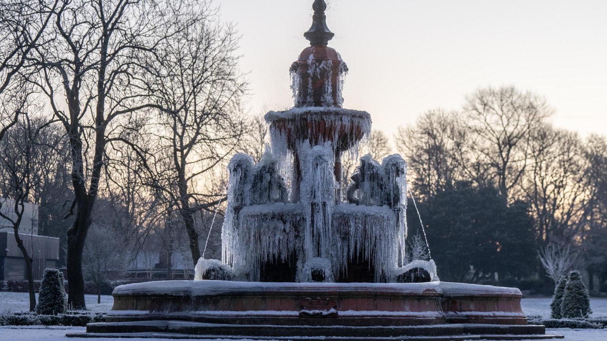 A large tiered water fountain stands in the middle of a frosty park. The fountain is covered in icicles hanging from every tier of the fountain. Behind the fountain are large trees. 