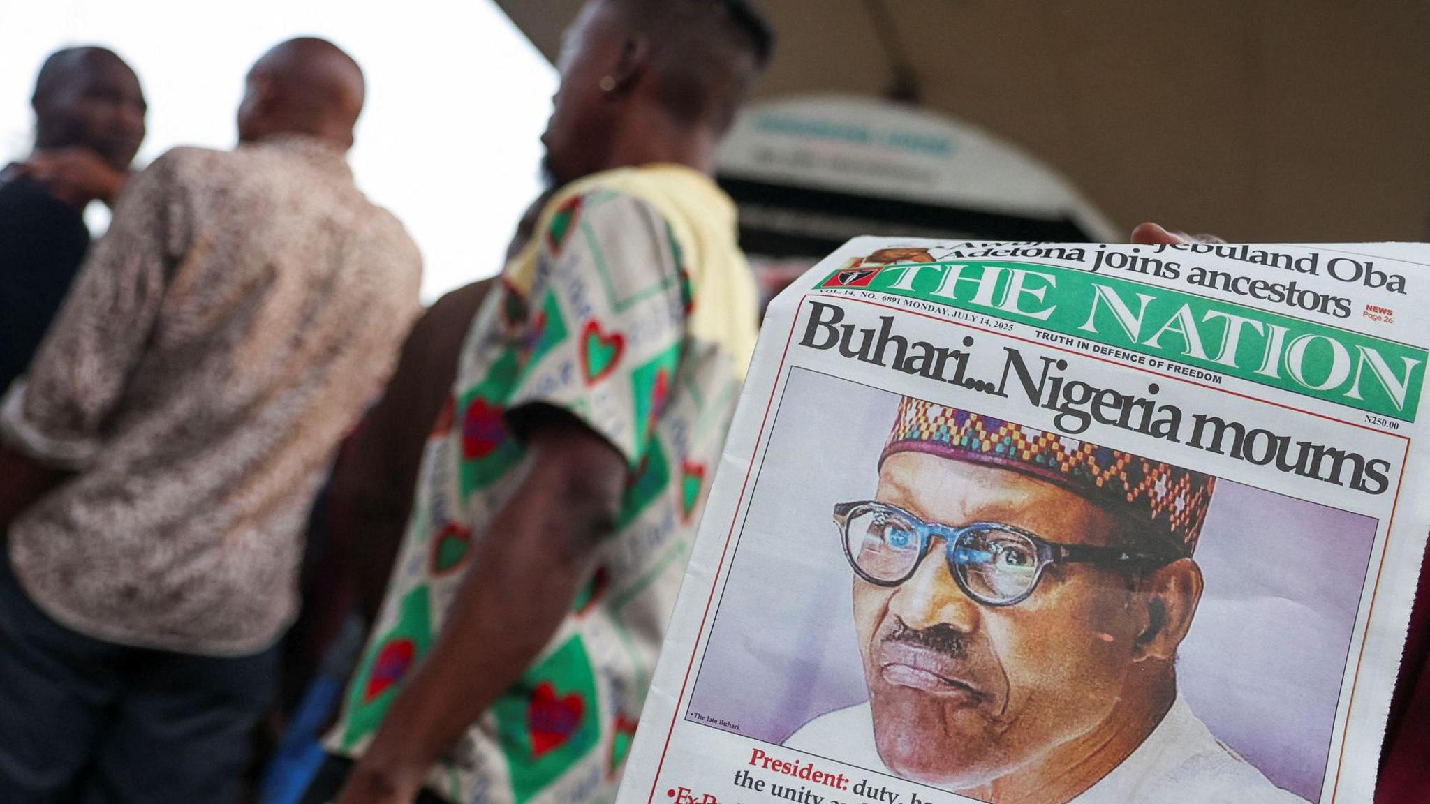 A person displays a newspaper carrying a headline reporting Muhammadu Buhari's death. Three men appear in the background of the image, out of focus.
