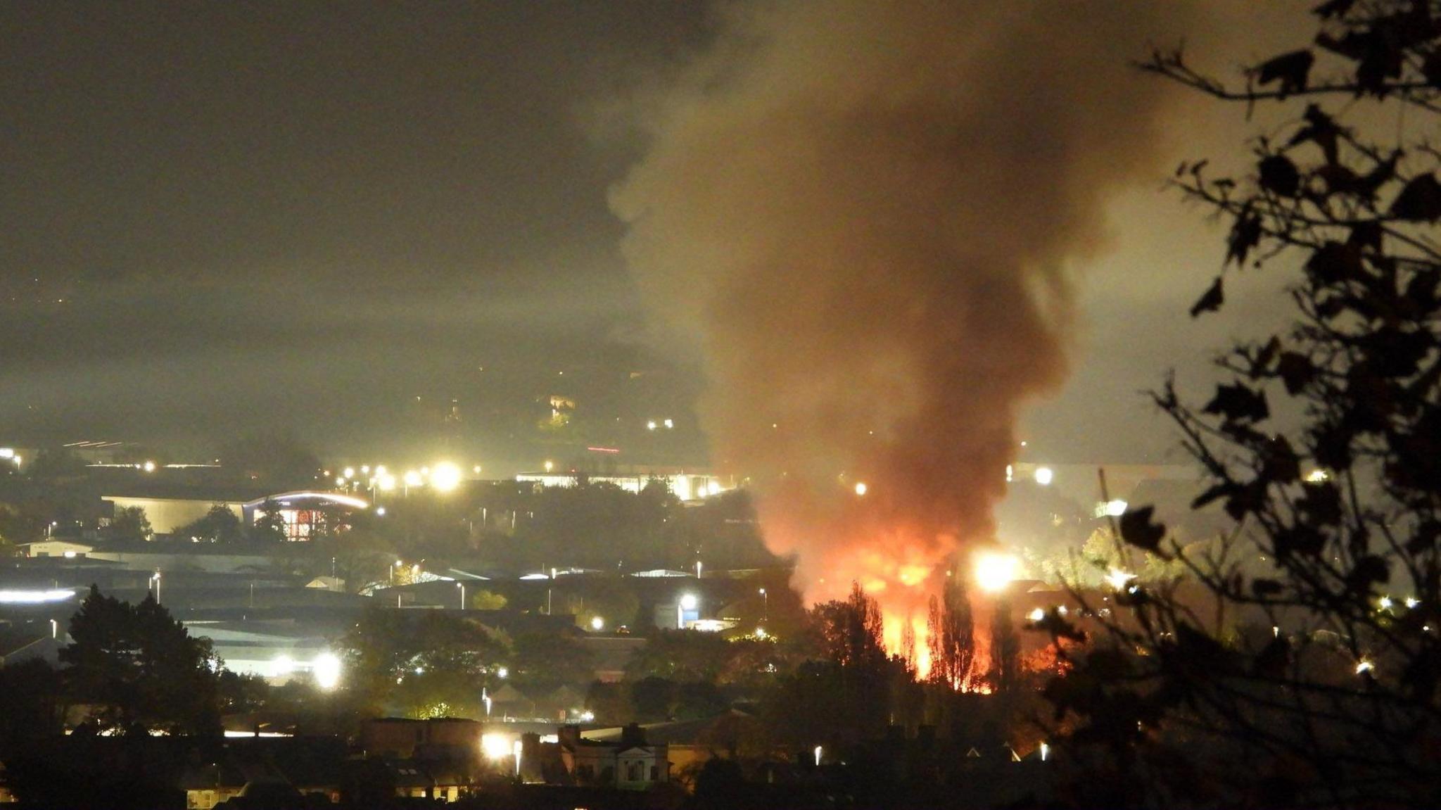 A large plume of smoke in the night sky at a distance. There are trees in the foreground. There is an orange glow from the flame at the bottom of the smoke. There are buildings surrounding the area.