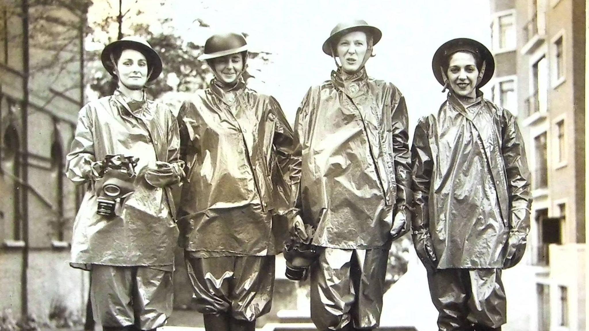 Four women standing, looking at the camera, wearing gas protection suits which appear as though made of tin foil