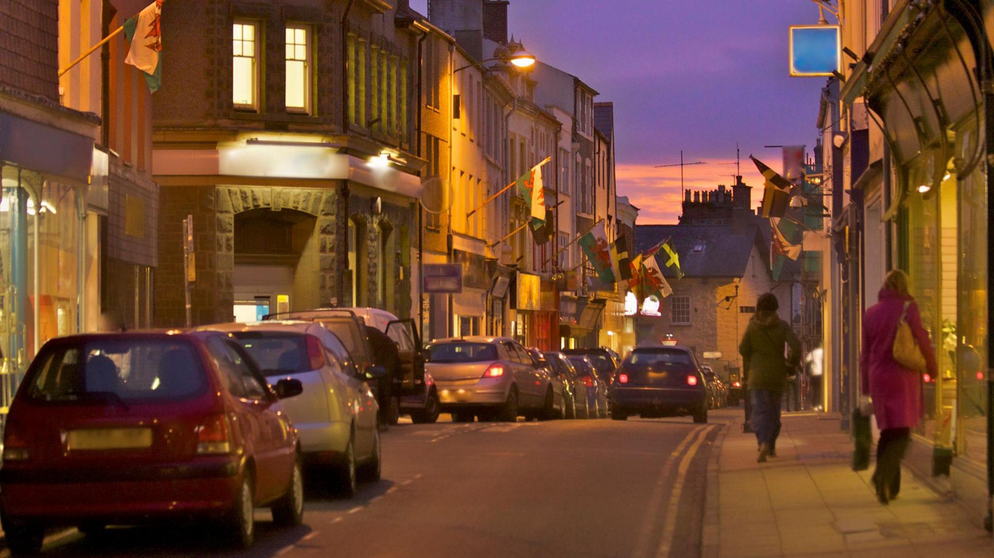 A picture of the town centre in Pwllheli. The sun is setting and therefore the ski is a purple colour. There are cars and people walking on the pavements.