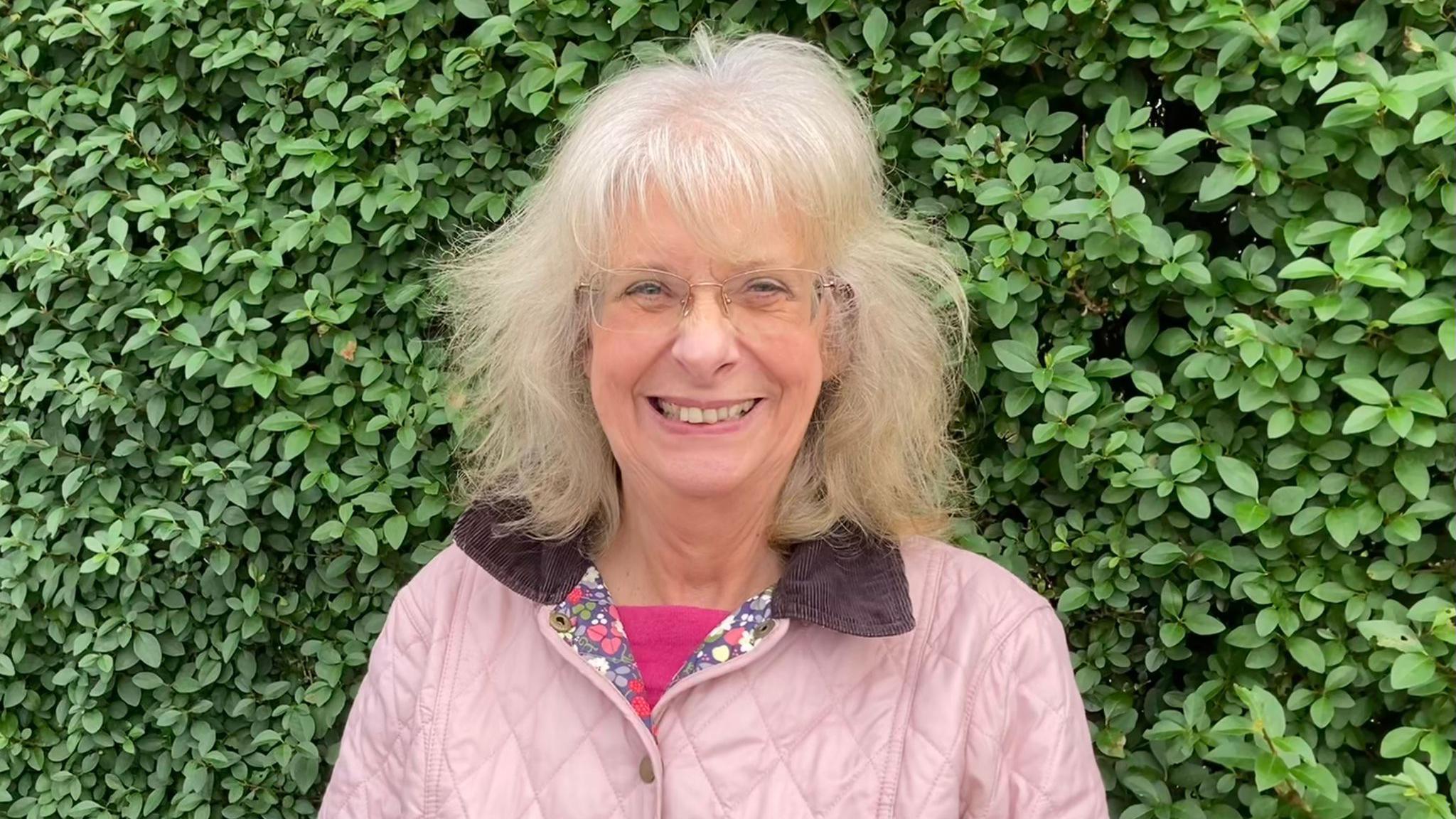 A woman smiling with rimless rectangle glasses on, a pink coat and a dark pink shirt underneath, with a bush behind her.