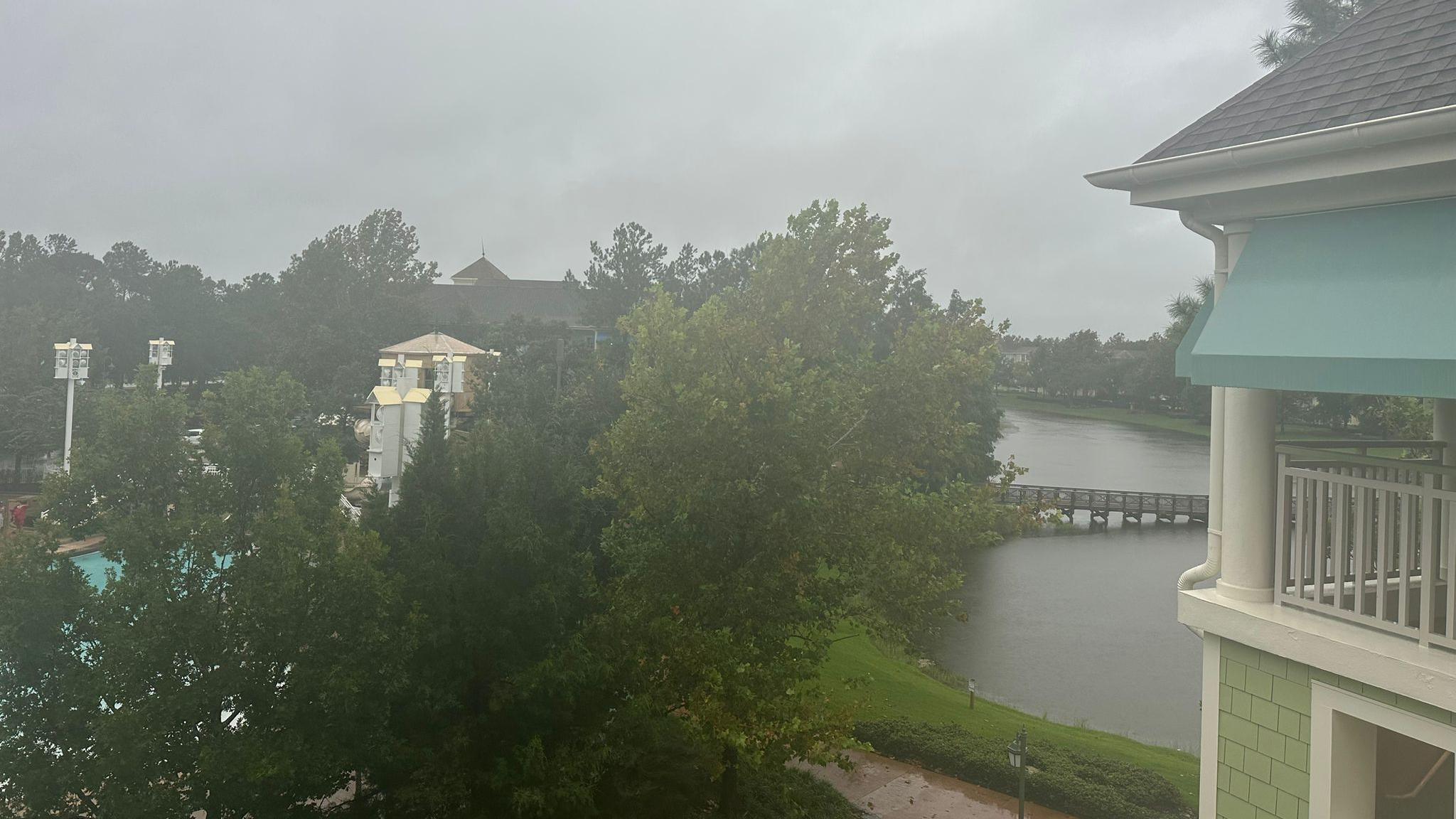 The scene outside the family's hotel window. There are trees and a lake. The clouds are grey, it is raining and it looks very stormy.