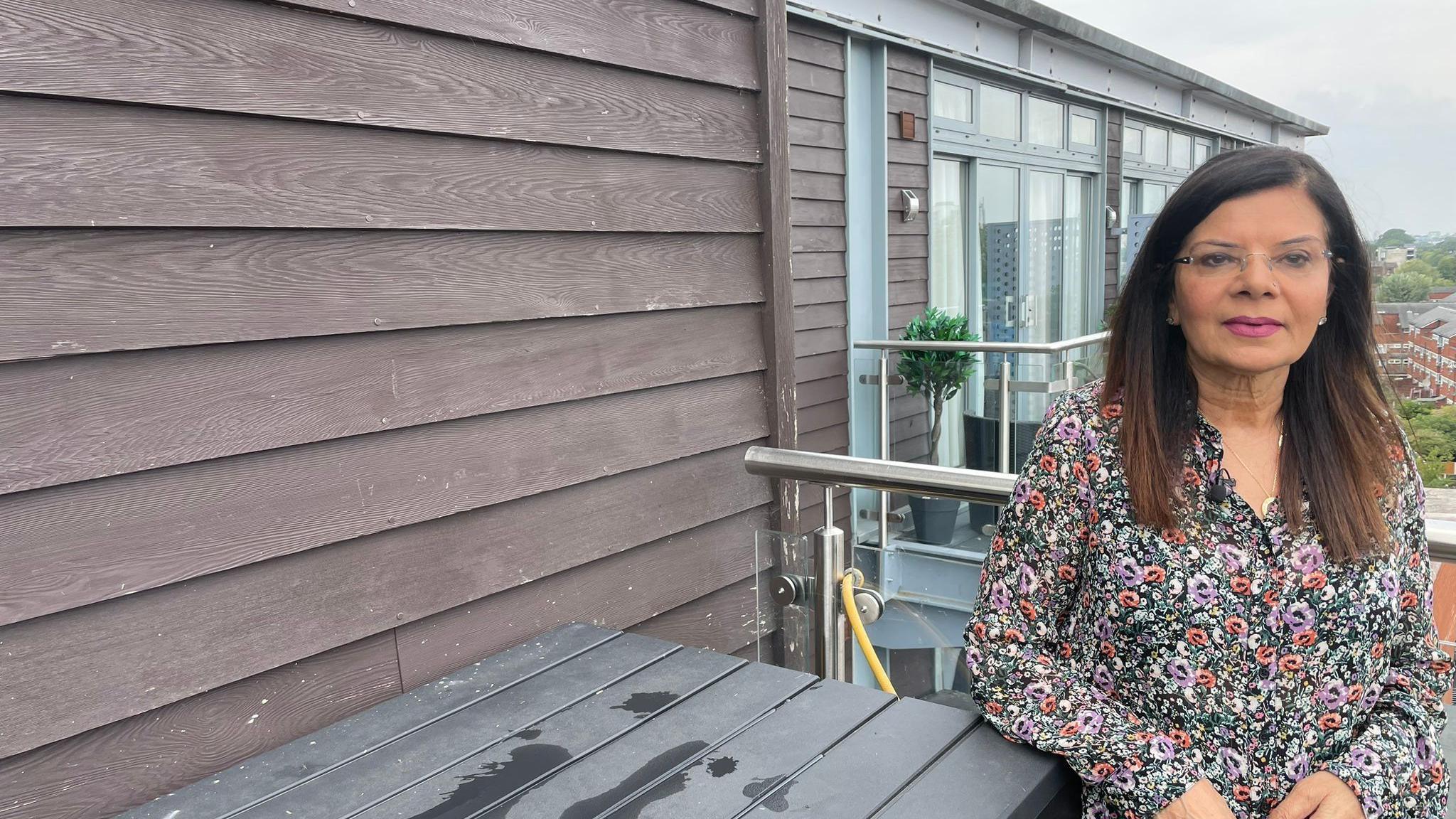 A woman in a floral dress stands on a flat balcony, with wooden panelling visible on the wall