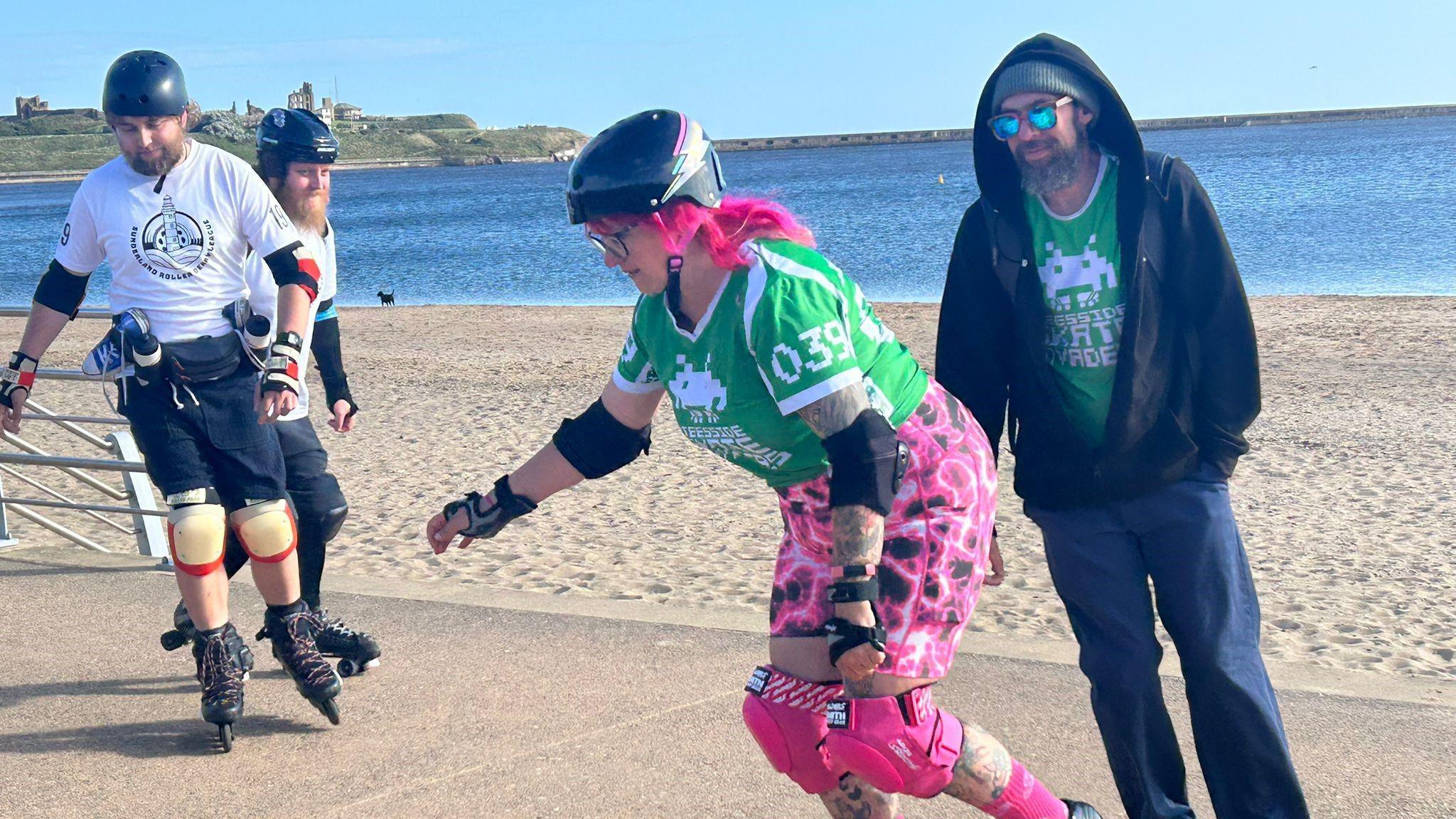 A group of people skating on a concrete area in front of the seafront. The woman in the foreground has pink hair and trousers with two men skating in the background. 