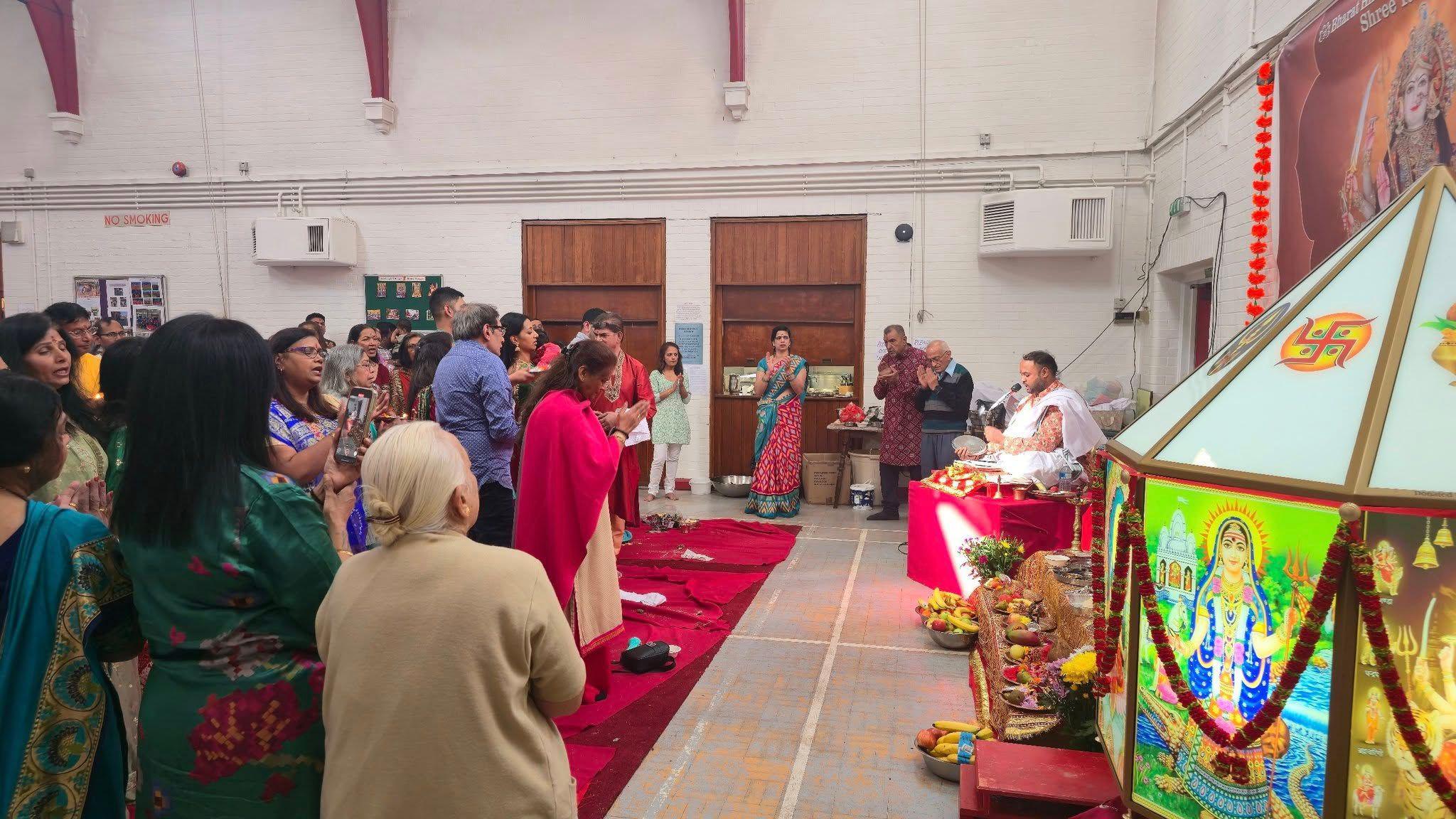 Members of the Hindu congregation standing in prayer. They are facing the priest in a white ceremonial outfit who is sitting next to pictures of Hindu gods.