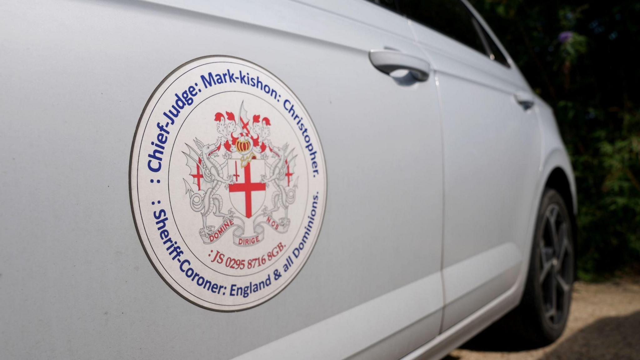 A white Volkswagen Polo with the crest of Mark Christopher on it. The crest has the St George cross in the middle with dragons either side.