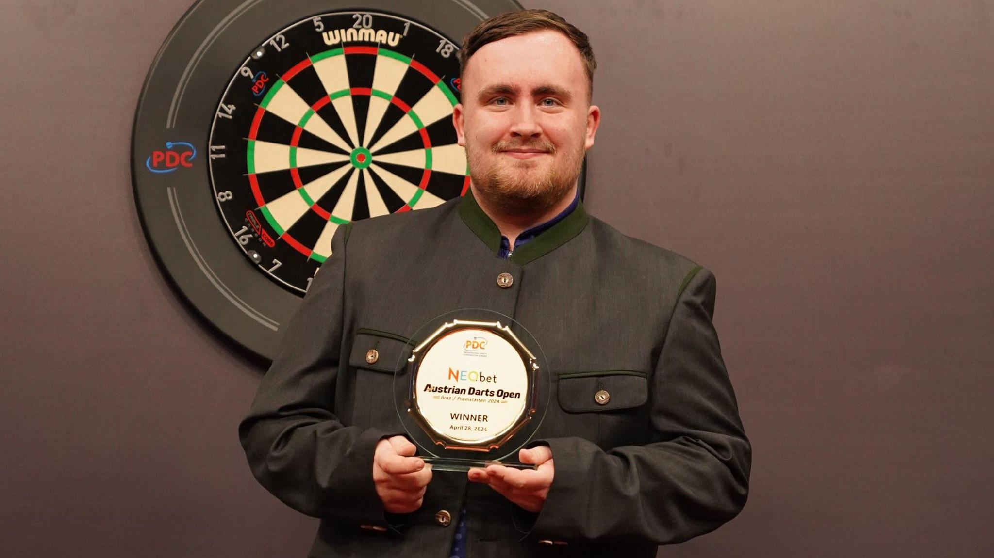 Luke Littler holds the Austrian Open trophy while wearing the winner's green jacket