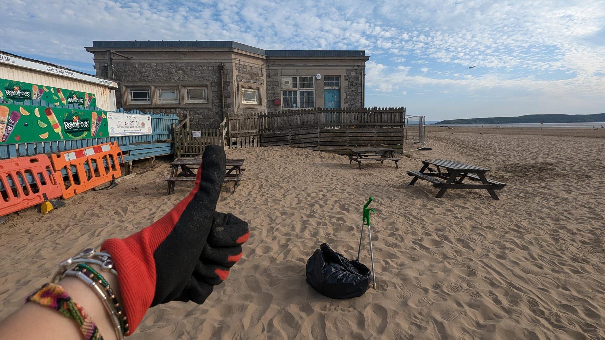 Ms Newton holding a thumbs up in front of the camera, wearing a black and red glove. Behind her hand there is a patch of sand which she has cleaned up, with a bin bag and litter picking tool visible