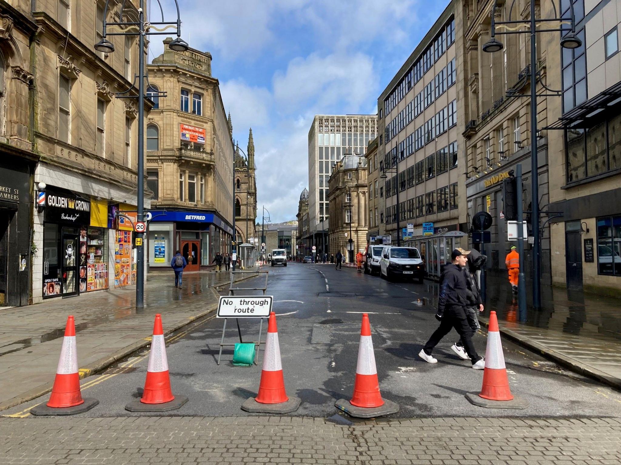 Five red and white traffic across a closed-off street with people walking across.