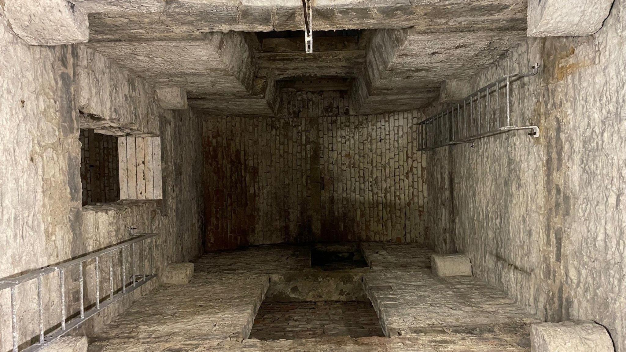 An underground stone passageway, deep and of some age. There are modern ladders on the left and right of the room. 