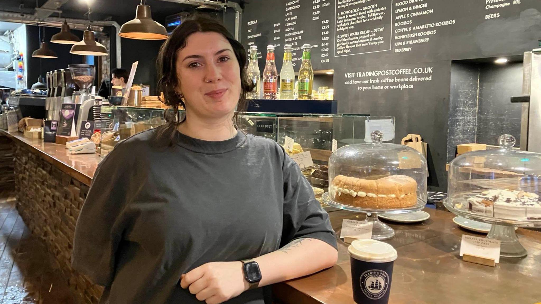 Ellen Davoudi-Meredith stands in front of the counter in a coffee shop with cakes under covers behind her and a menu printed on a blackboard behind her. She is wearing a grey t-shirt and has a coffee cup next to her.