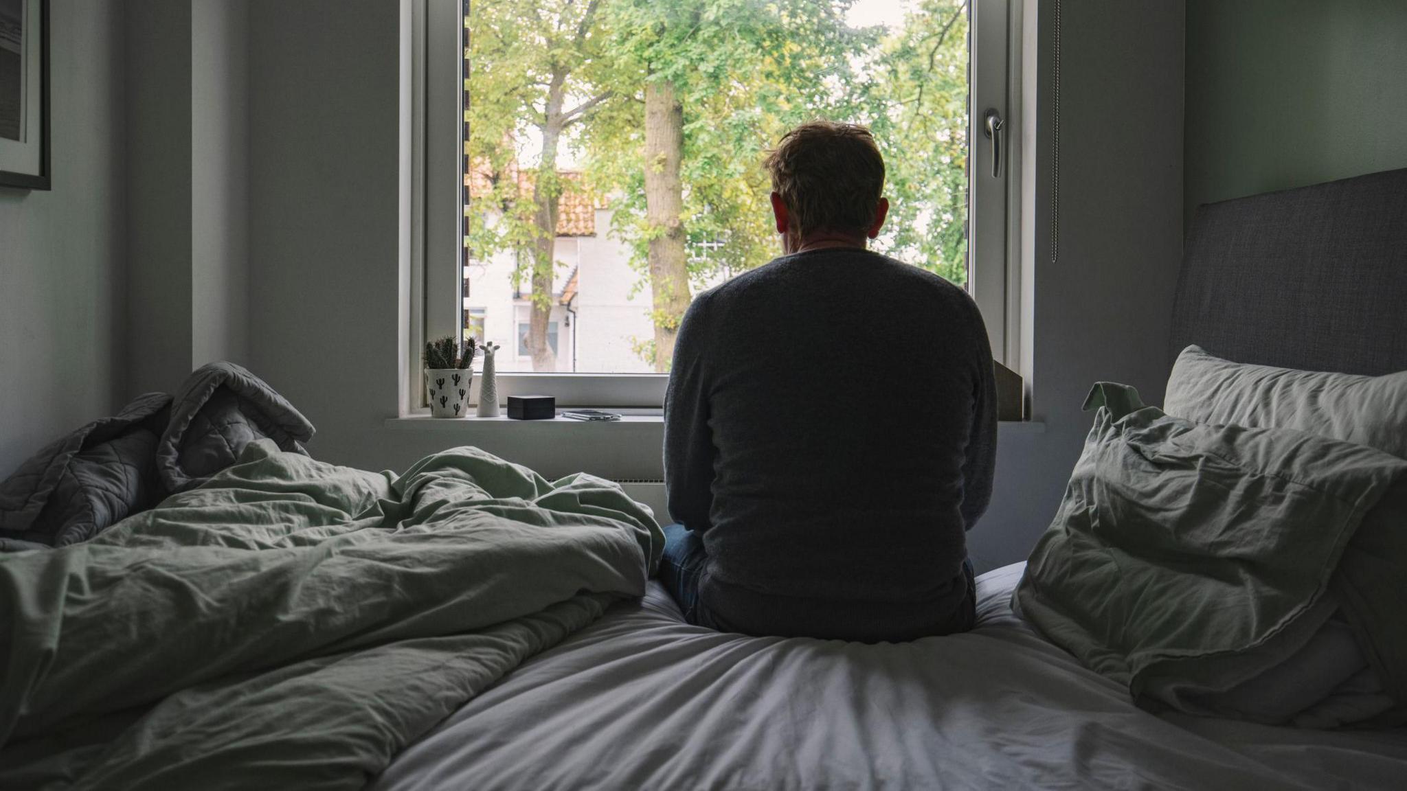 A man sits on a bed with green and grey sheets, seen from behind, looking out of a window towards trees outside.