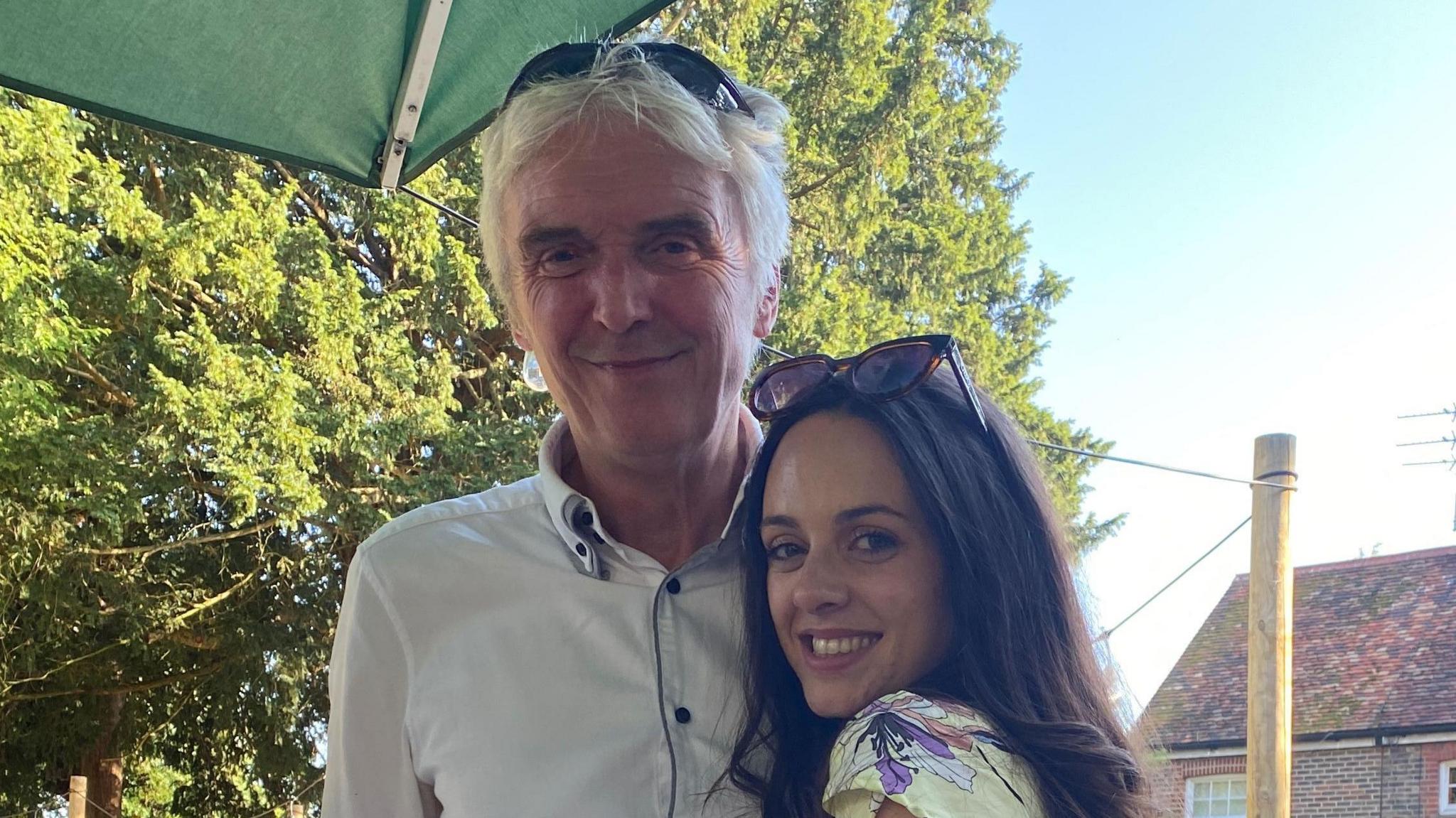 A man with short white hair wearing a white shirt and sunglasses on his head. He is stood next to his daughter, who has brown hair and is wearing a floral top and sunglasses on her head.