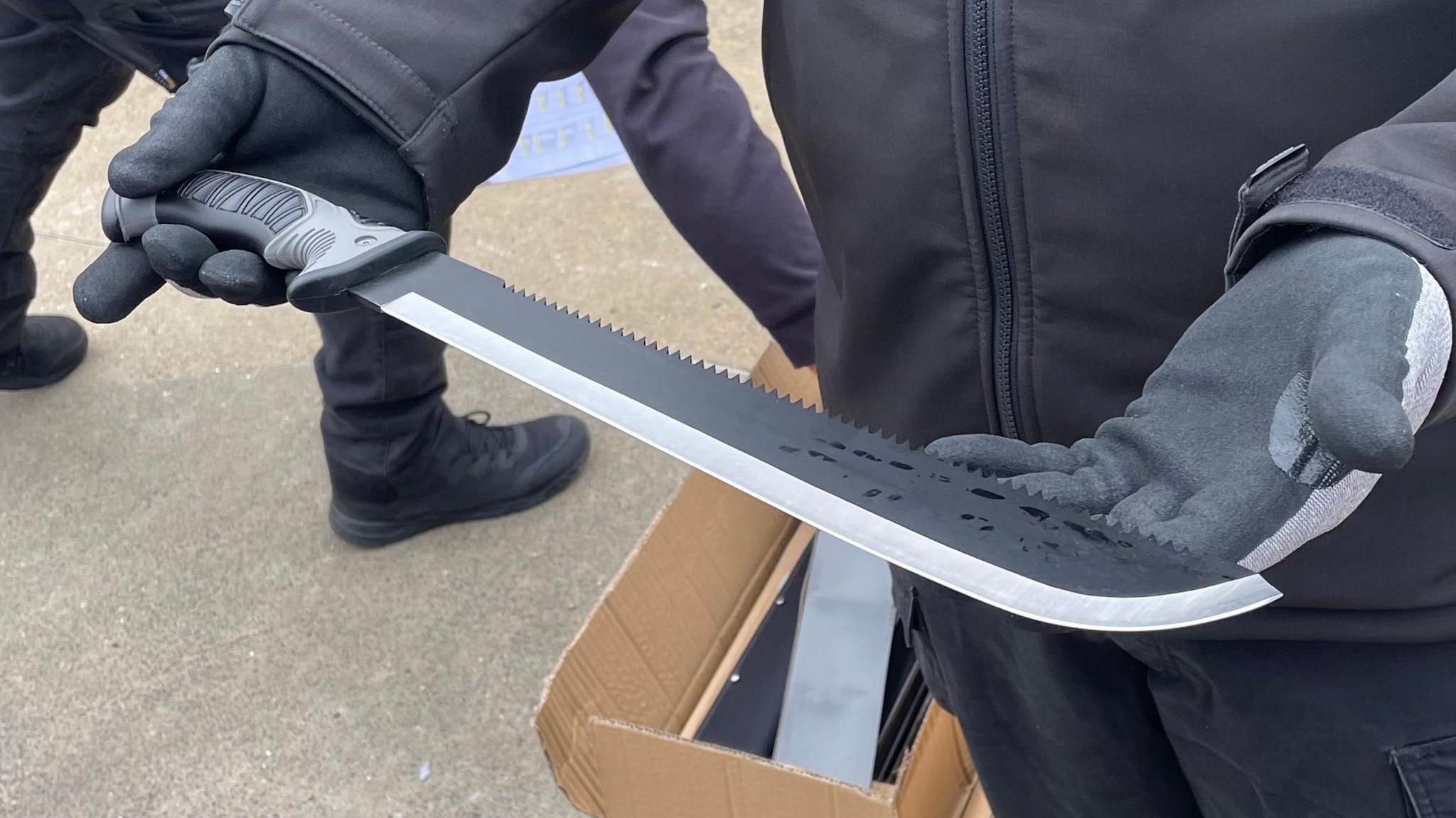 A large knife with a blade that is sharpened on one side and serrated on the other. It is being delicately held by two hands wearing heavy-duty black safety gloves. In the background is a cardboard box containing smaller black boxes; it is sitting on a concrete surface. Another person wearing black boots and trousers appears to be reaching into the box. 