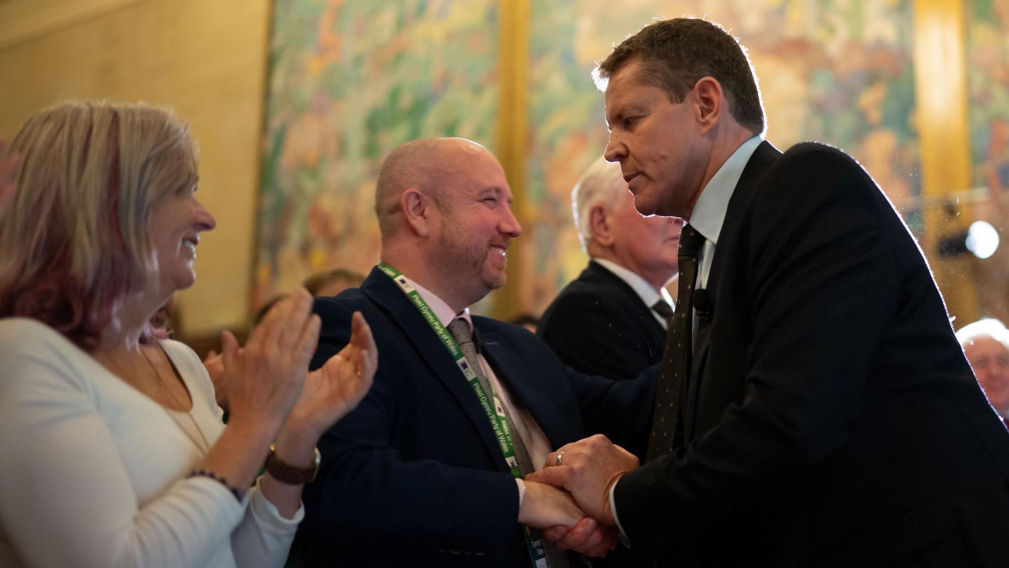Liz Saville Roberts clapping to the left in a white top, with Mabon ap Gwynfor holding Rhun ap Iorwerth's hands in the centre of the picture. Both Ap Gwynfor and Ap Iorwerth are wearing black suits.