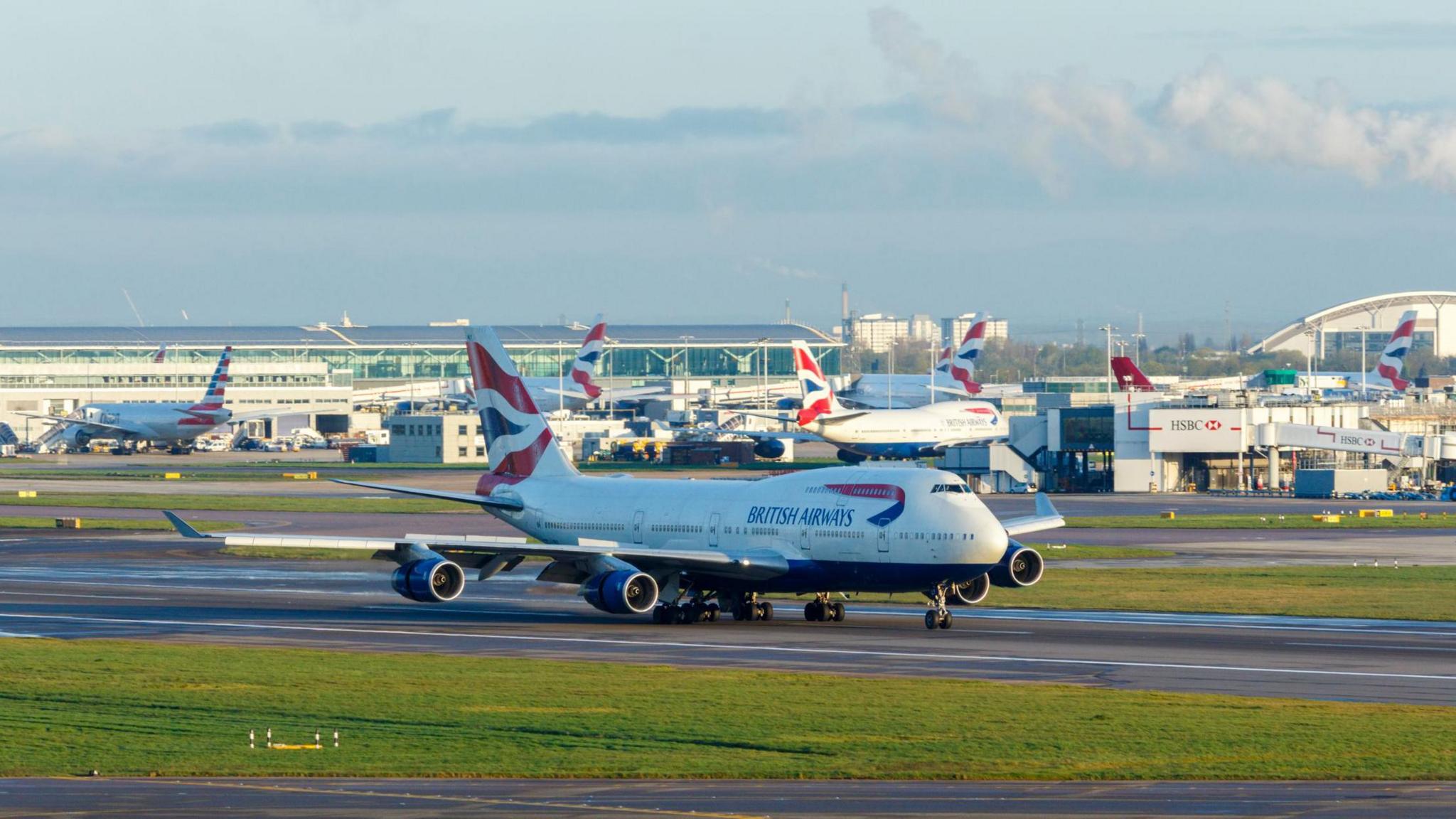 A British Airways plan is seen taxiing at Heathrow Airport. Other planes and airport facilities are seen in the background.