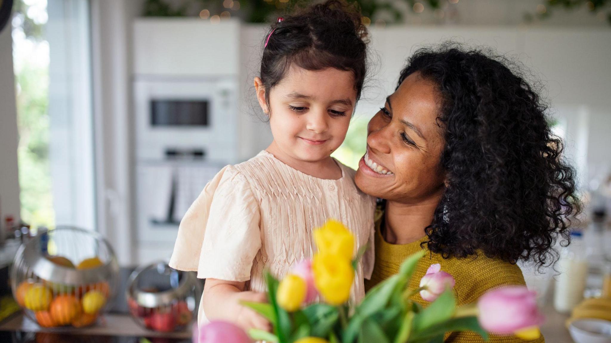 A girl wearing a pale pink dress is held by a woman in a mustard top with curly hair. A vase of yellow and pink tulips and a basket of oranges and lemons are in the foreground.