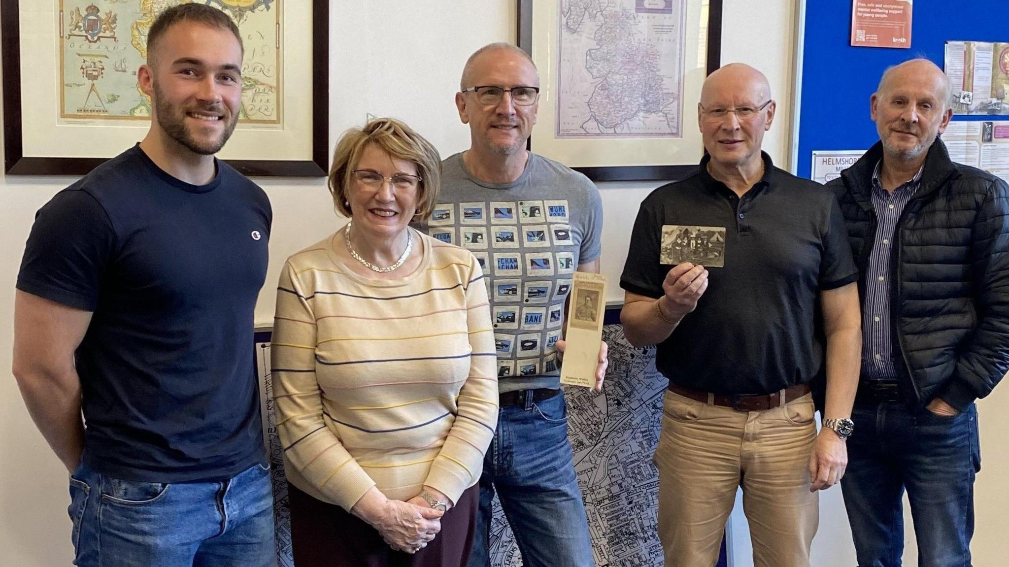 A young man in his 20s wears a blue t-shirt and jeans, standing next to one elderly woman and three older men at a library. Two men in the middle are holding WW1 postcards