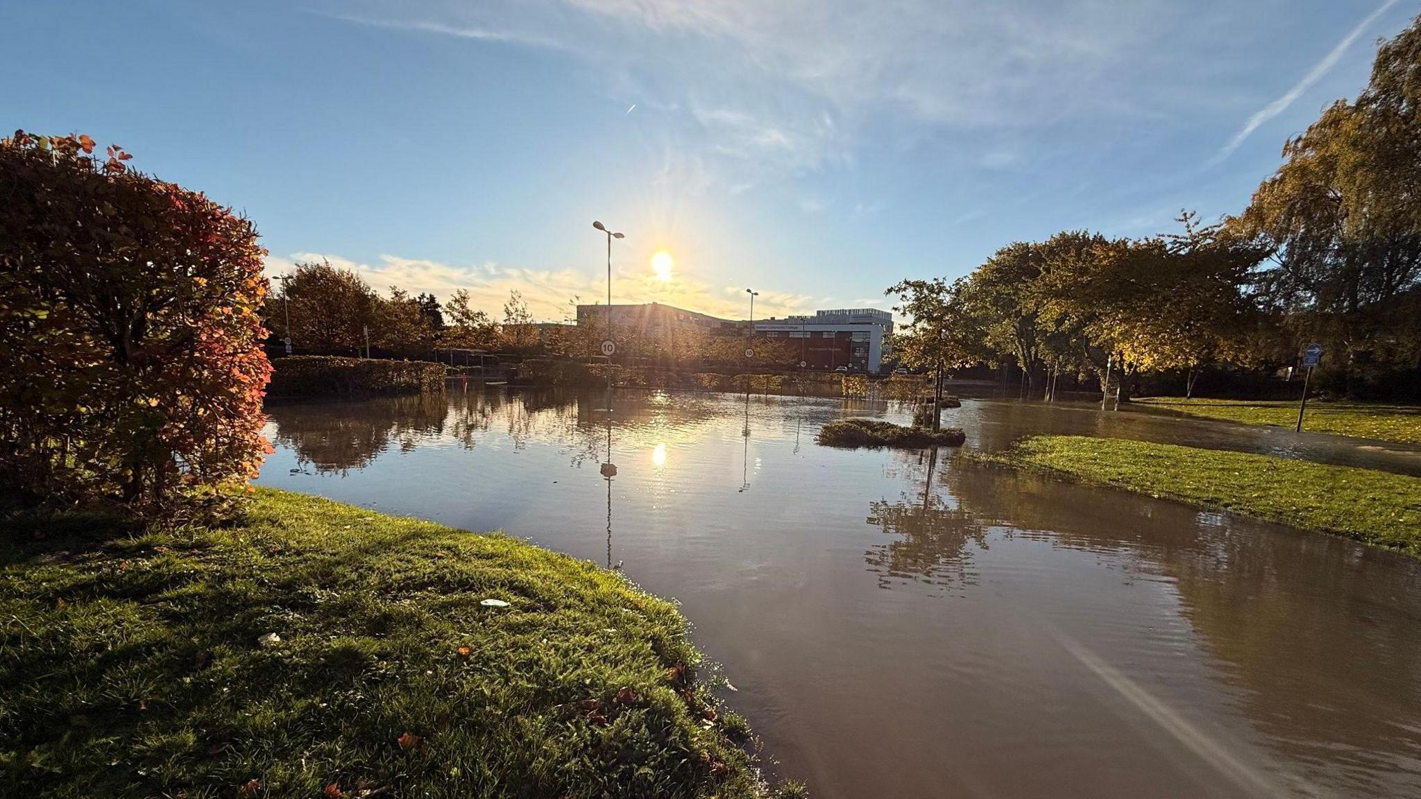A flooded outdoor area with water covering a road and grassy sections, reflecting trees and lampposts in the sunlight. The sun is low in the sky, casting a warm glow over autumn-colored foliage on the left and trees on the right. A building is visible in the background under a partly cloudy blue sky.