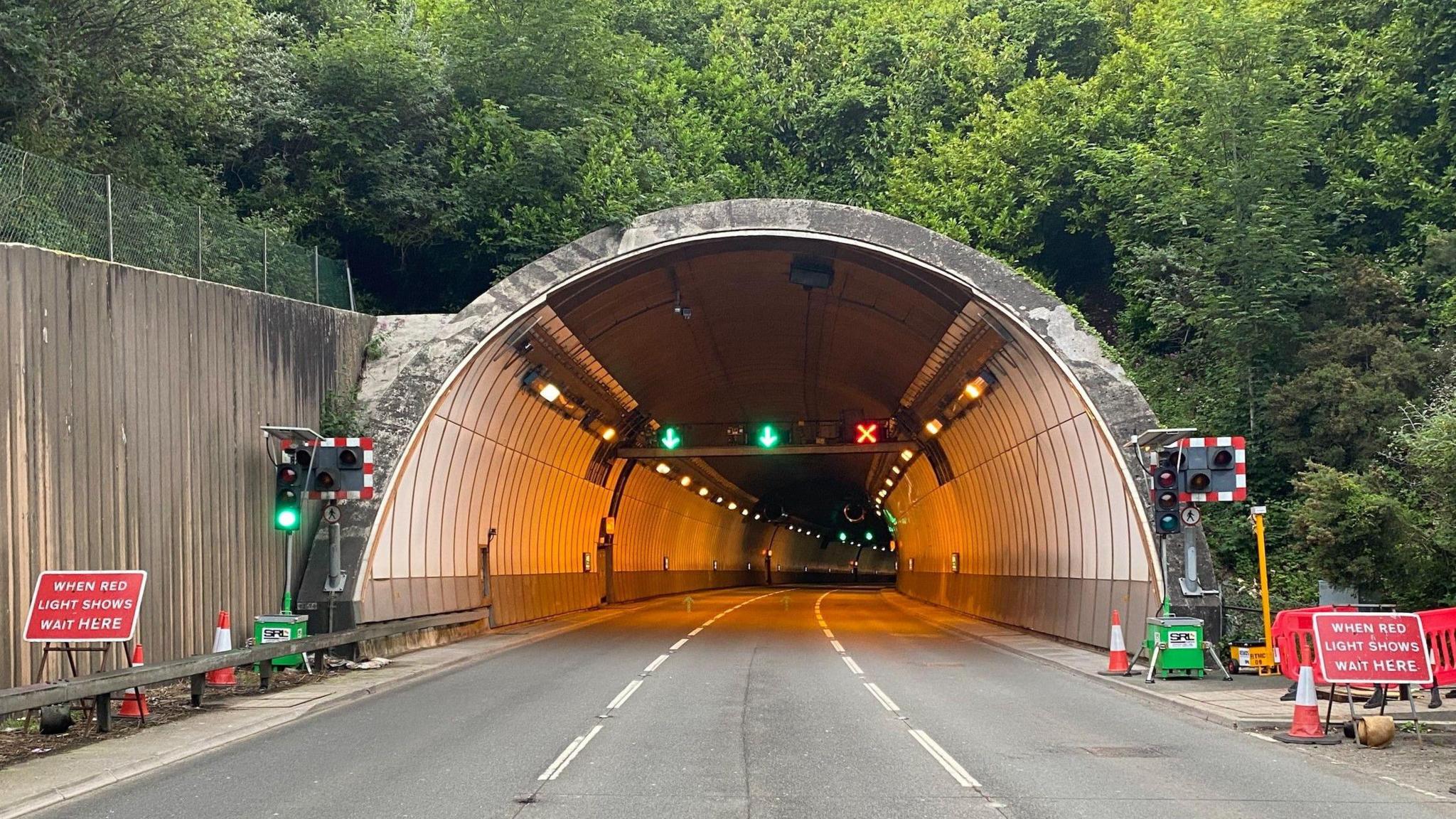 Saltash Tunnel with traffic lights and signs at the side of the tunnel. There are two green arrows inside the tunnel on the left and red cross on the left. There are green trees surrounding the tunnel.