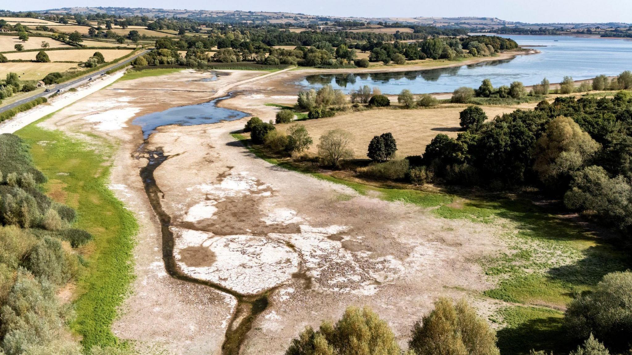 A picture taken from a drone showing the extent of the receding water of Chew Valley Lake near Bristol. Large dry patches with small creeks running through them are visible, some baked white in the sun, where there would normally be water. Some water can be seen in the middle of the lake in the distance. A main road is seen passing the lake and there are some low hills with trees and pale grass patches on them.