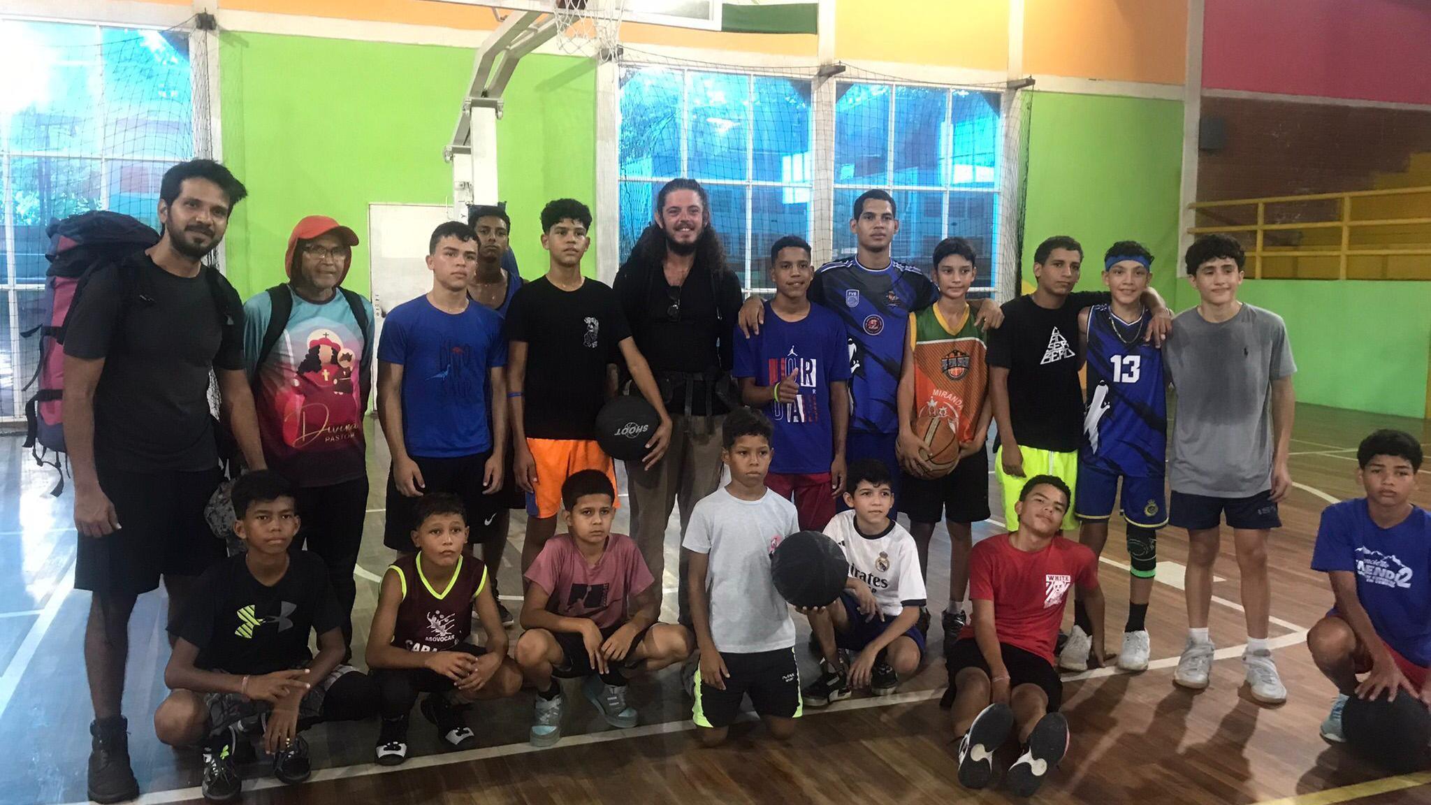 Ollie Treviso is in a school gymnasium with a group of predominantly teenage boys who are in two rows. The back row is standing and the front row are sitting down. Some of them are holding basketballs. The walls of the gym are multicoloured.
