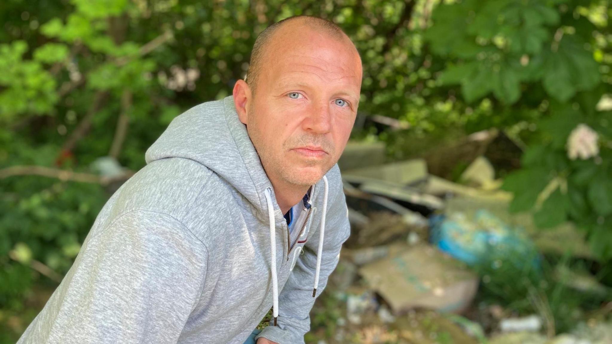 A man with shaved hair and a grey hoodie squats down next to piles of rubbish in woodlands