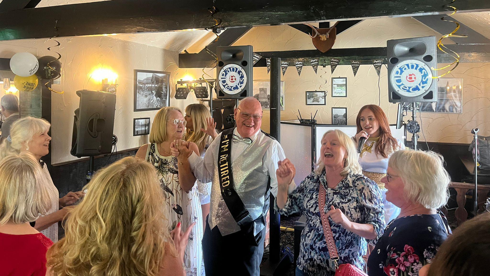 A bald man with glasses is wearing a sash that reads "I'm retired", he is holding hands with two women who are singing and mid-dance. Behind them is an auburn haired woman who is singing into a microphone