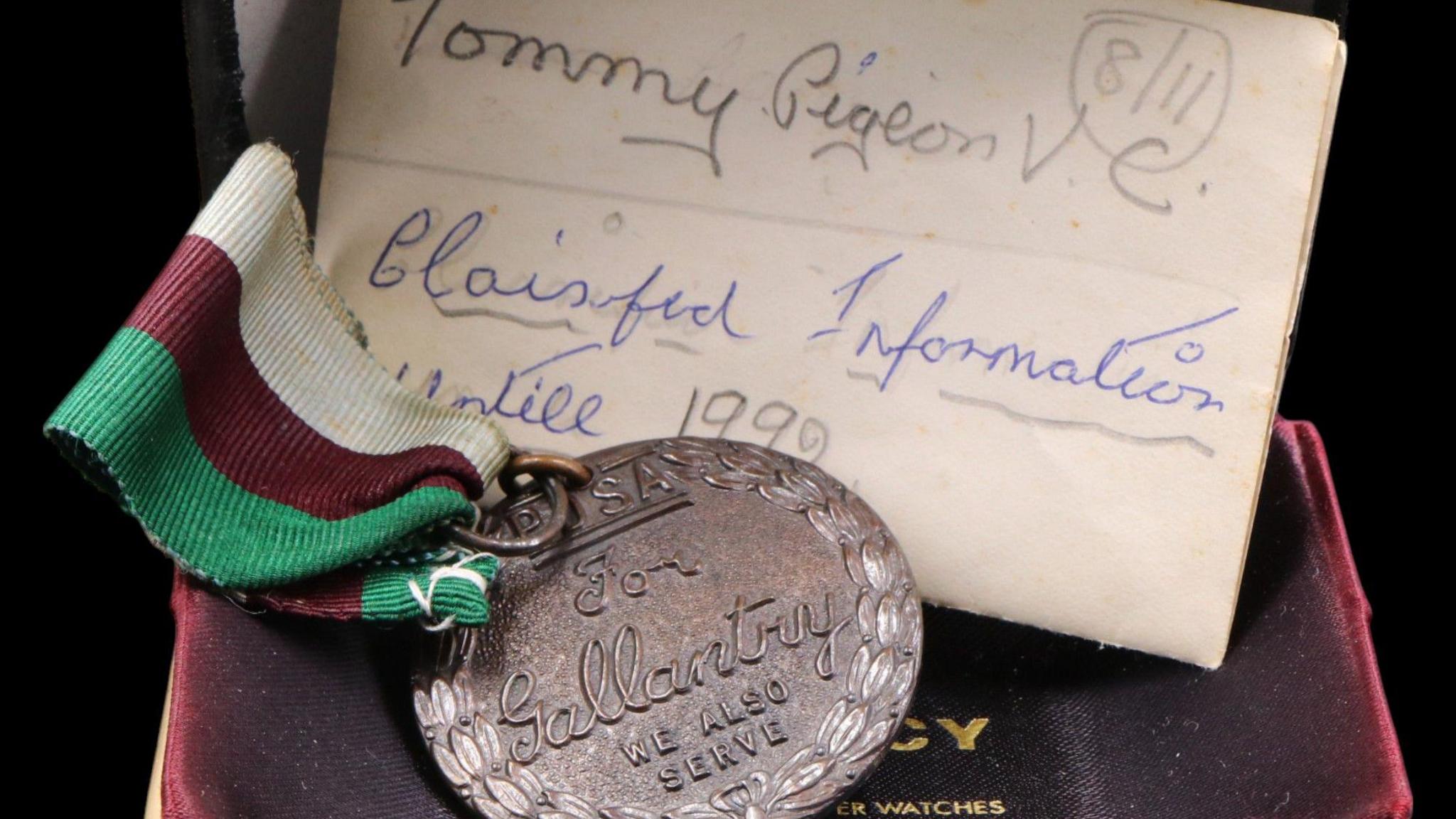 The Dickin Medal awarded to Tommy is shown in a red velvet box. It has a red, green and white ribbon and there is a piece of paper tucked behind it.