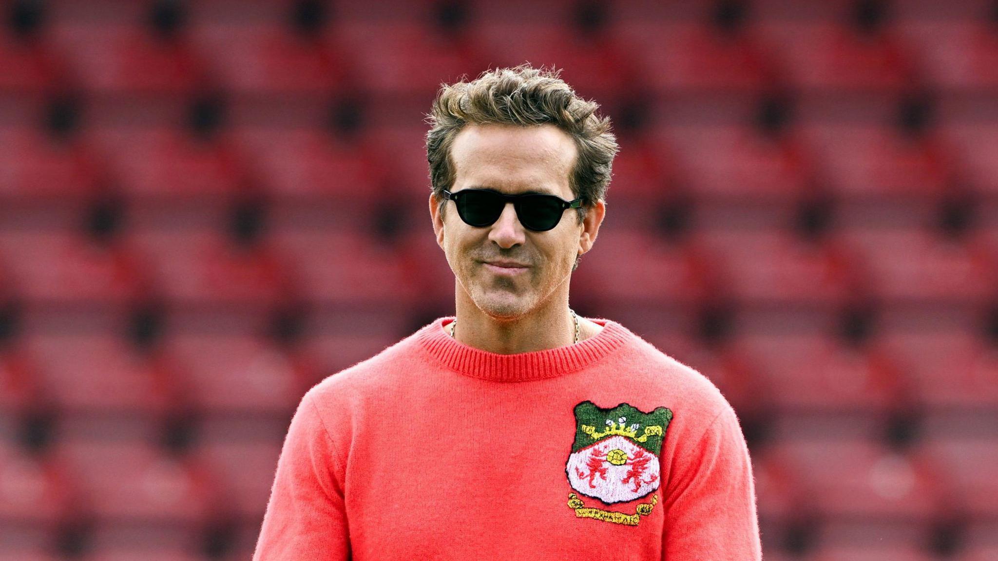 Actor Ryan Reynolds wearing a red Wrexham AFC knitted jumper and beige trousers. He is walking across the pitch at the football stadium with the empty red seats visible in the background. He is smiling and wearing dark lens sunglasses.