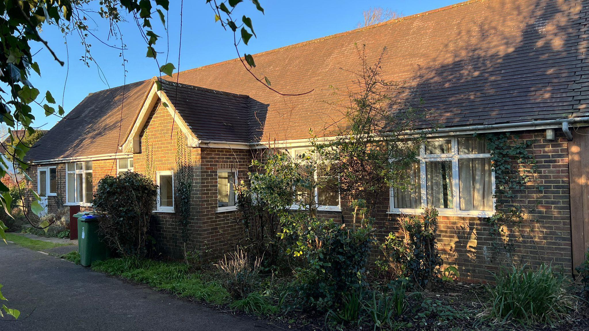 A single floor, long, brown brick building with white wooden windows along the front. It's triangular roof is also brown. there are messy overgrown plants in the foreground. The sky behind the building is blue