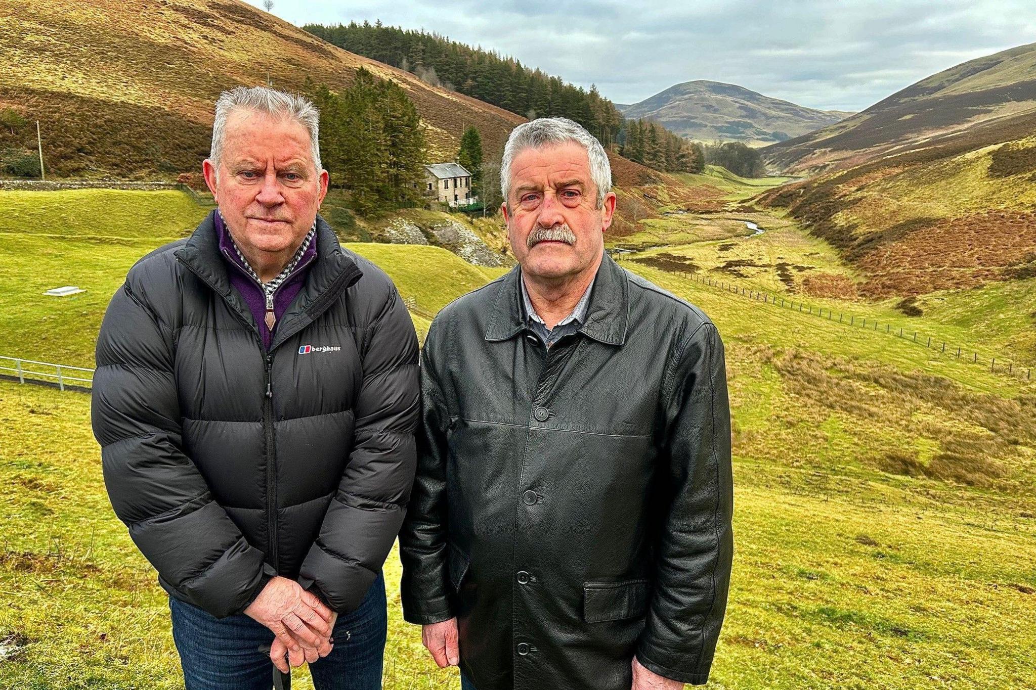 Bill is on the left in a black puffa jacket. He has dark blue jeans on and a walking stick. He has grey hair. There is a green valley and hills behind him. Dougie is on the right and is wearing a black leather jacket. He has grey hair and a moustache.