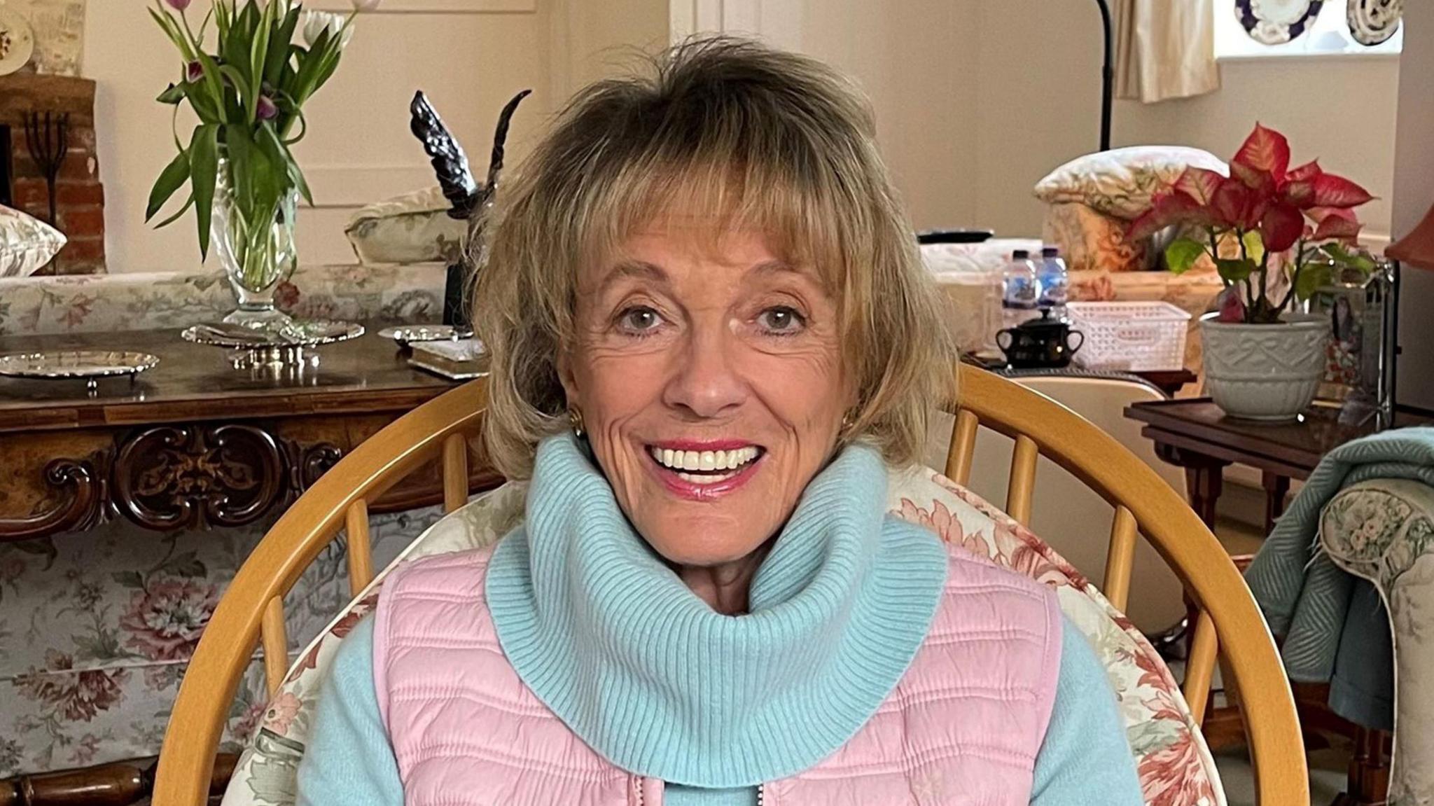 Dame Esther Rantzen sits in a wooden backed chair with a red floral print cushion for her back to rest on. She is smiling at the camera, has short blonde hair with a fringe framing her face and wears a pale blue roll-neck sweater and pink gilet. Behind her is a living room, with a pink and white floral print sofa, ornate wooden console table with a glass vase holding tulips.