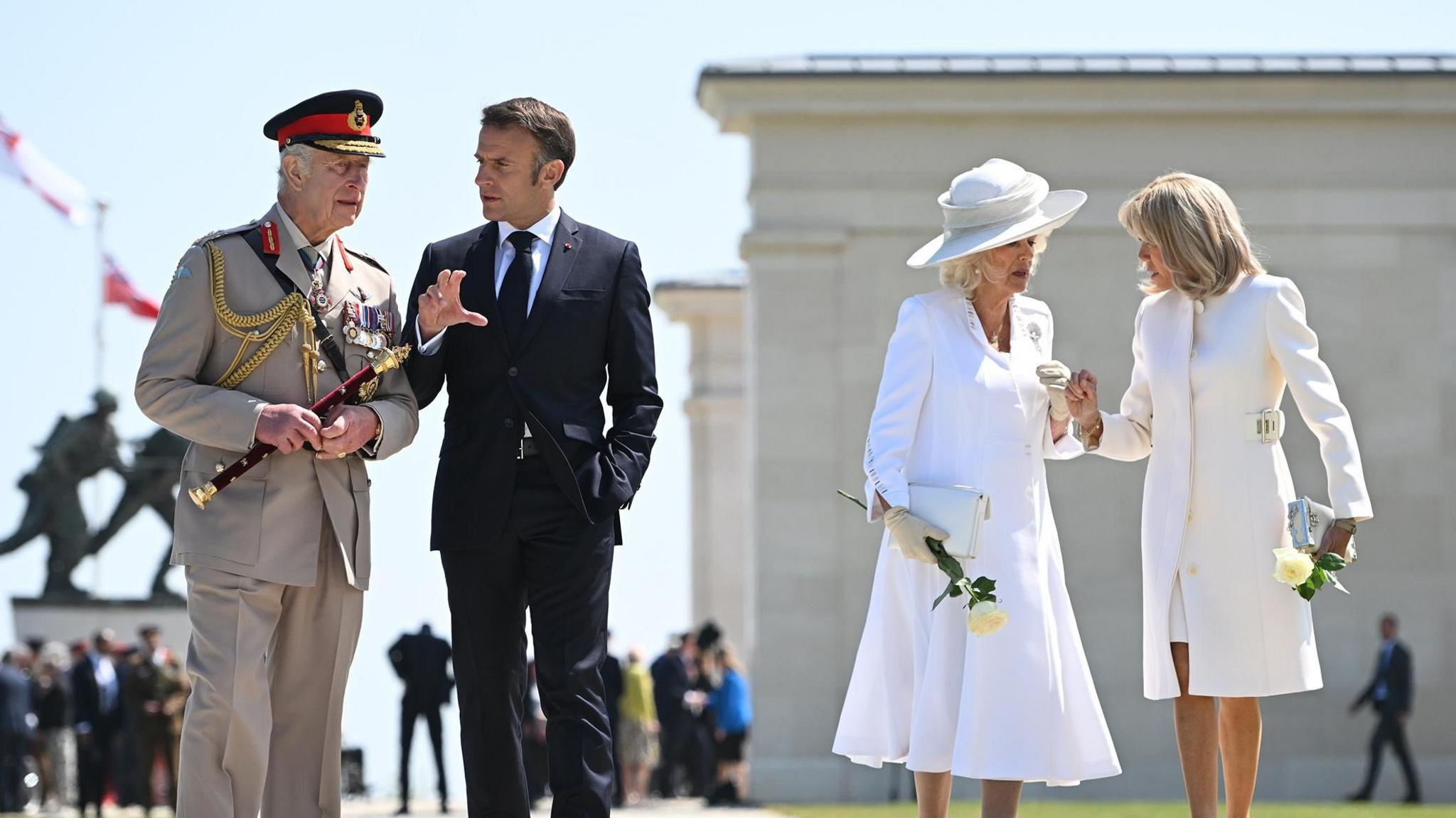 King Charles and French President Emmanuel Macron talk and walk together. Behind and to the side of them, Queen Camilla and Brigitte Macron do the same. Behind them is the British Normandy Memorial in Ver-sur-Mer, France.