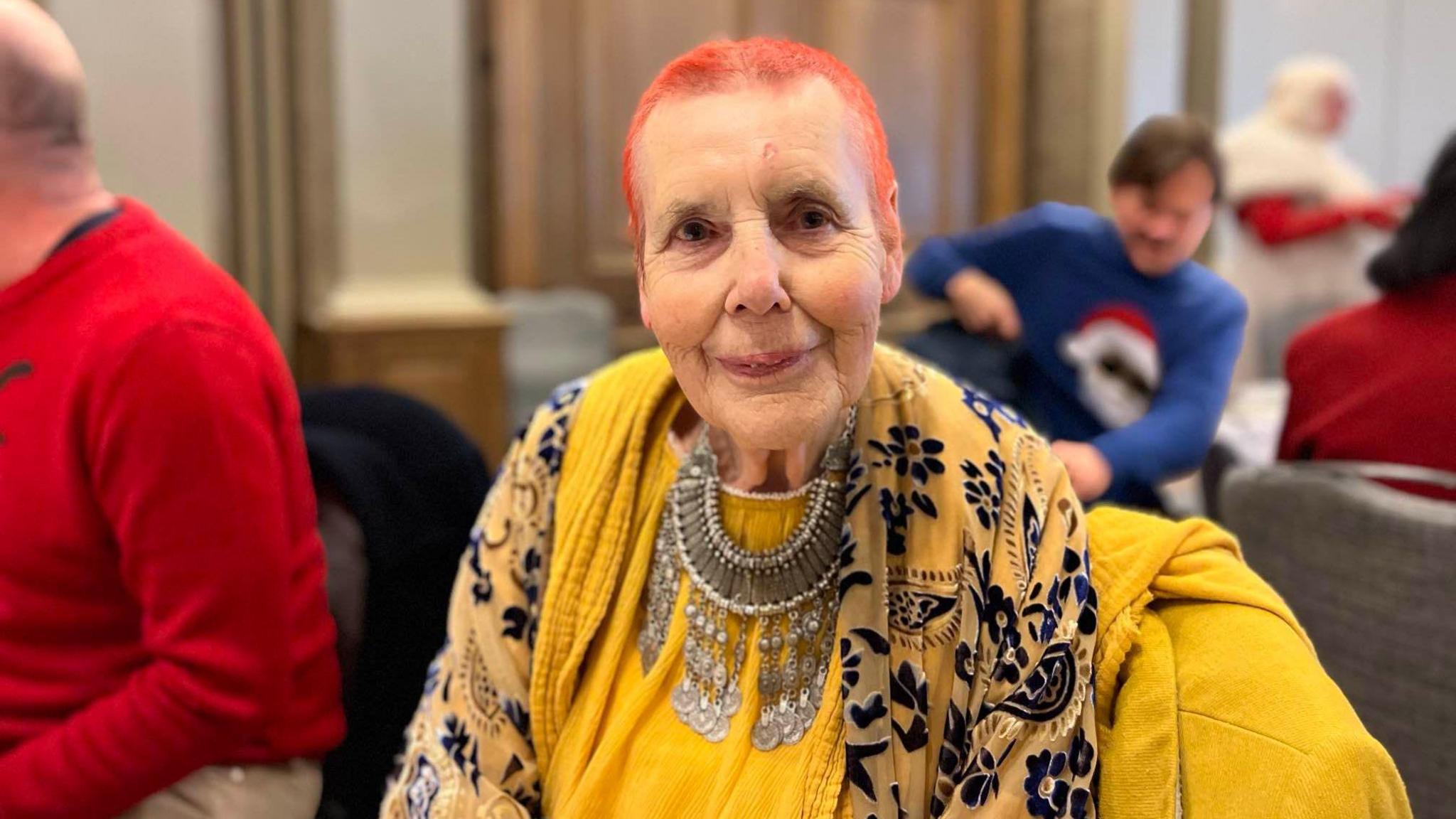 Hilary Howard at the festive party. She is wearing a bright yellow top, yellow jacket and a large necklace. She has orange hair and is looking directly at the camera. 