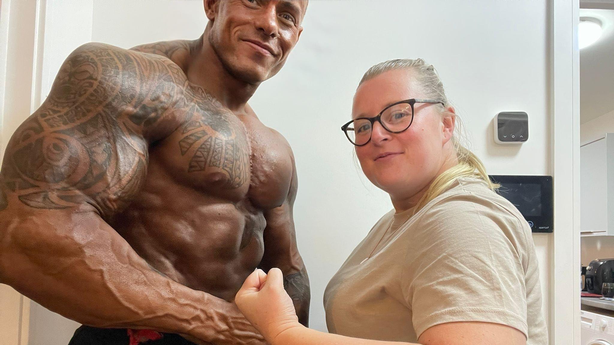 Jay Baggaley is smiling and posing while standing next to his partner. She is smiling with her fist clenched while looking at the camera. She has long blonde hair and is wearing a cream coloured T-shirt and glasses. 