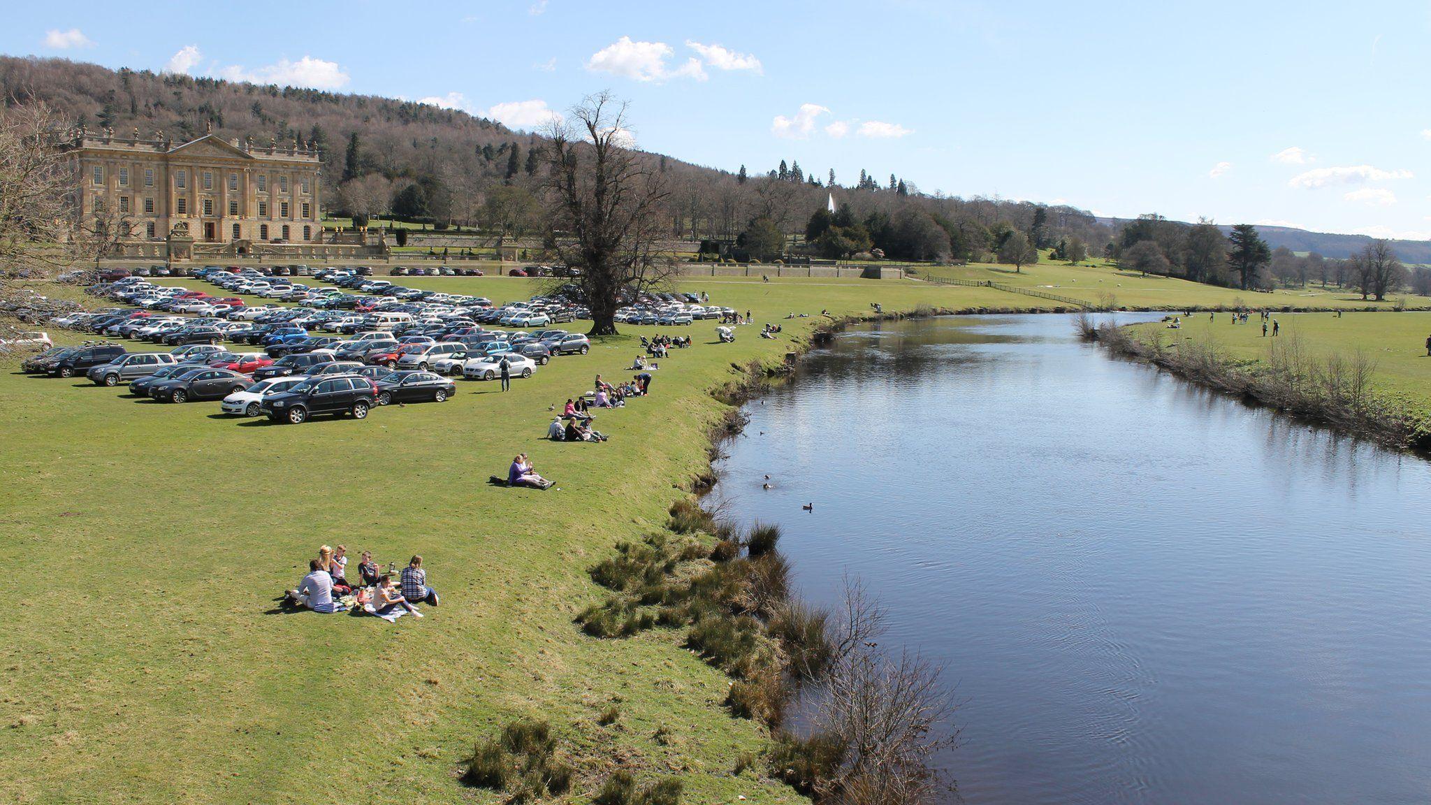 People sitting on a river bank with Chatsworth House visible in the background