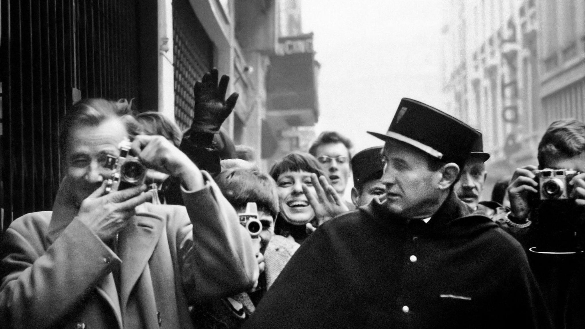 Titled: Photographers, fans, and officers, rue de Caumartin, Paris, January 1964. A black and white image of people looking at the camera, some are smiling, others are pointing cameras at the photographer, McCartney.