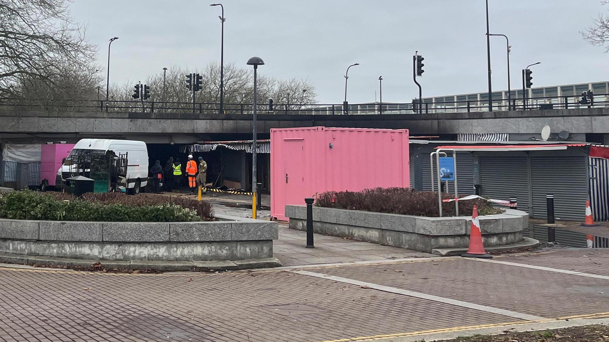 Some market stalls which have clearly suffered fire damage under a road bridge. There are other market stalls with their shutters down. A white van and a pink container can be seen behind some bollards and a small hedge in a concrete bed.