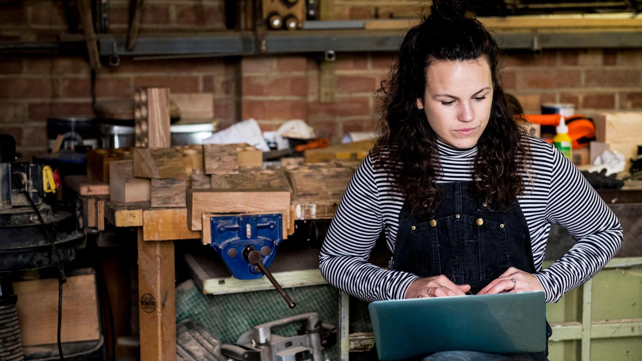 Young woman wearing dungarees and a stripey long sleeved top looking at a laptop in a carpentry workshop