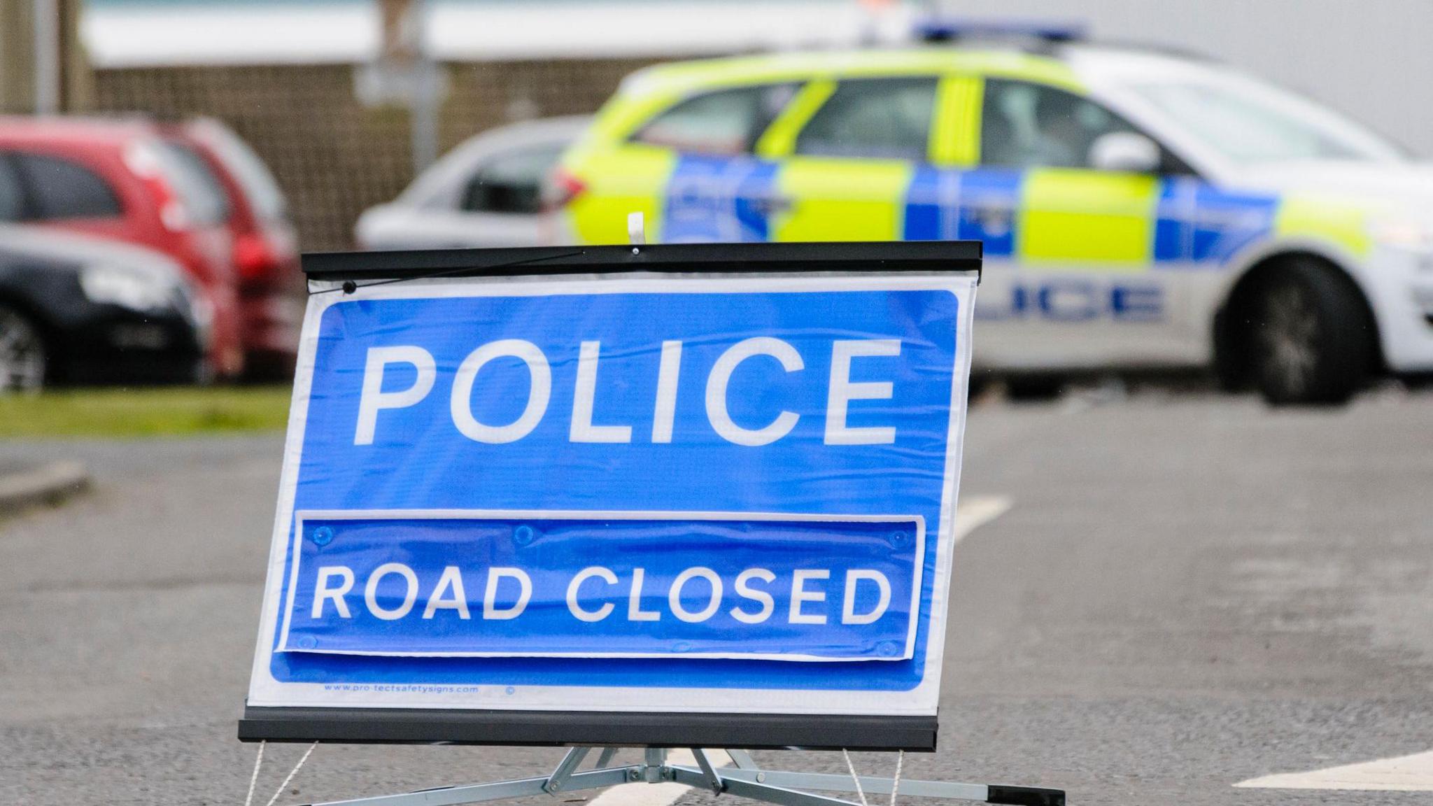 A blue police road closed sign with police car in background