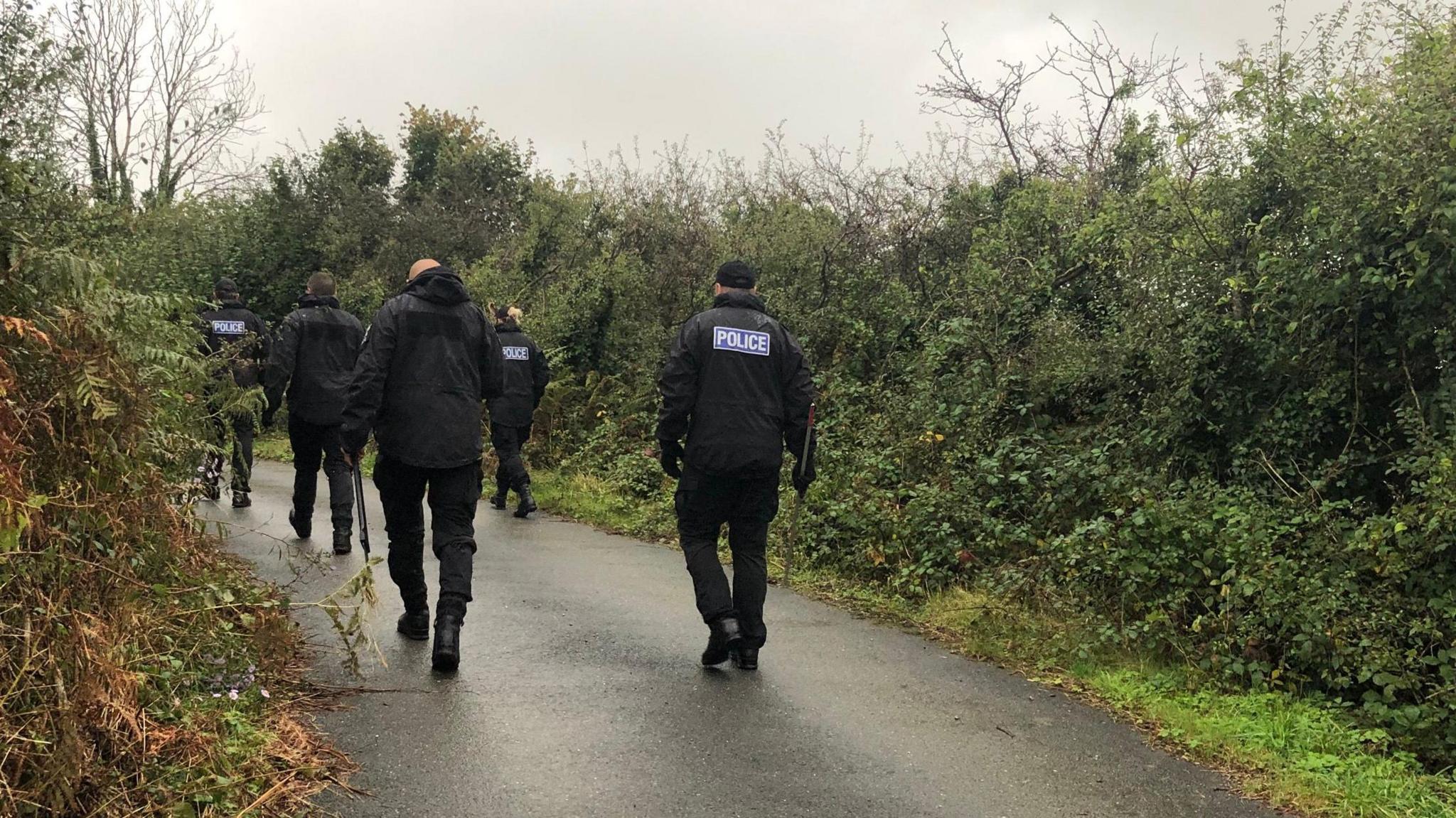 Police officers wearing black coats, some with the 'police' logo on the back, walking along a Cornish lane bordered by hedgerows